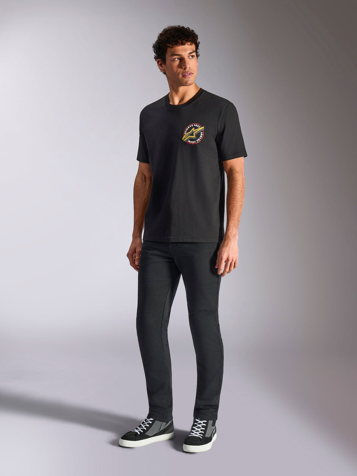 Alpinestars Aorbit CSF Tee, kortärmad t-shirt, svart, med en cirkulär Always Fast Made to Last-grafik på bröstet, bärs av en modell med mörka smala byxor och sneakers, casual motorsport-streetwear