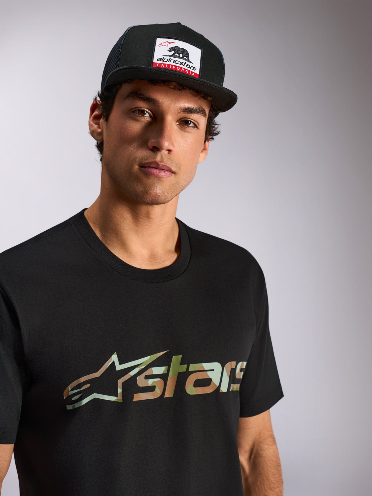 Alpinestars Verdure Camo CSF Tee, kortärmad t-shirt, svart, kamouflage-fylld Astars-logga, bärs av manlig modell med svart Alpinestars California trucker-keps, vardaglig motorsport-streetwear
