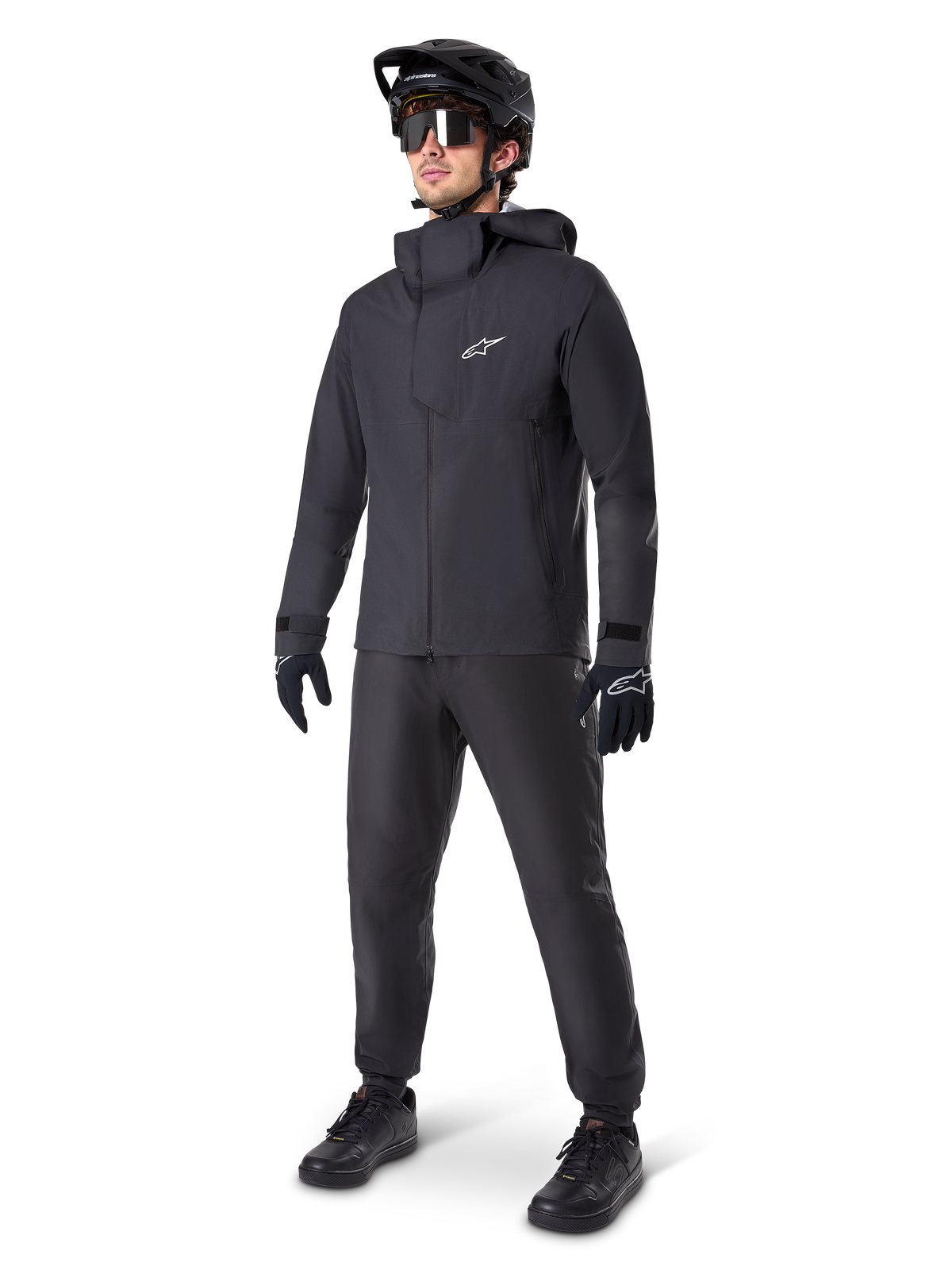 Alpinestars A-Dura Elite vattentät jacka i svart, bärs av en manlig cyklist med matchande svarta MTB-byxor, handskar och hjälm. Jackan har en teknisk vattentät design med en vit Astars-logotyp på bröstet och en hög skyddande krage.