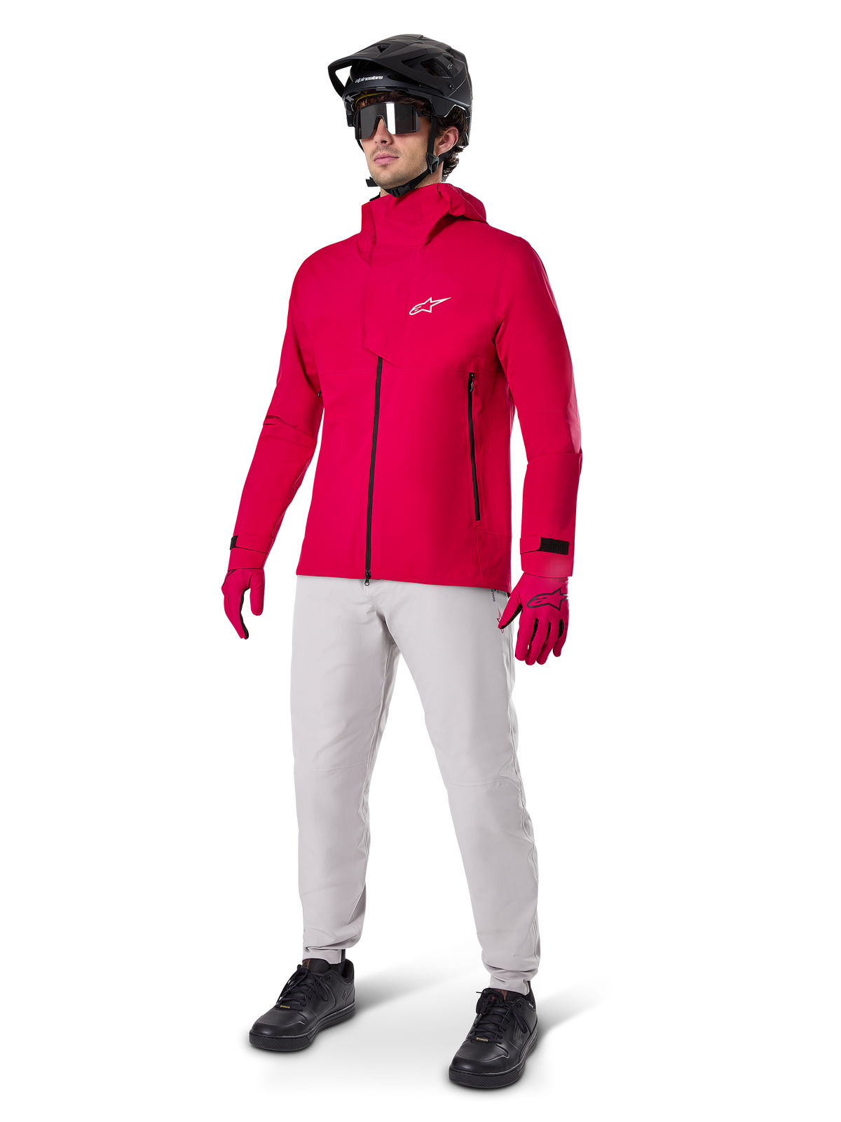 Manlig cyklist i Alpinestars A-Dura Elite vattentät jacka i Red Fluo, kombinerad med ljusgrå tekniska byxor, röda handskar, svart MTB-hjälm med goggles och svarta skor, komplett högpresterande outfit för mountainbike