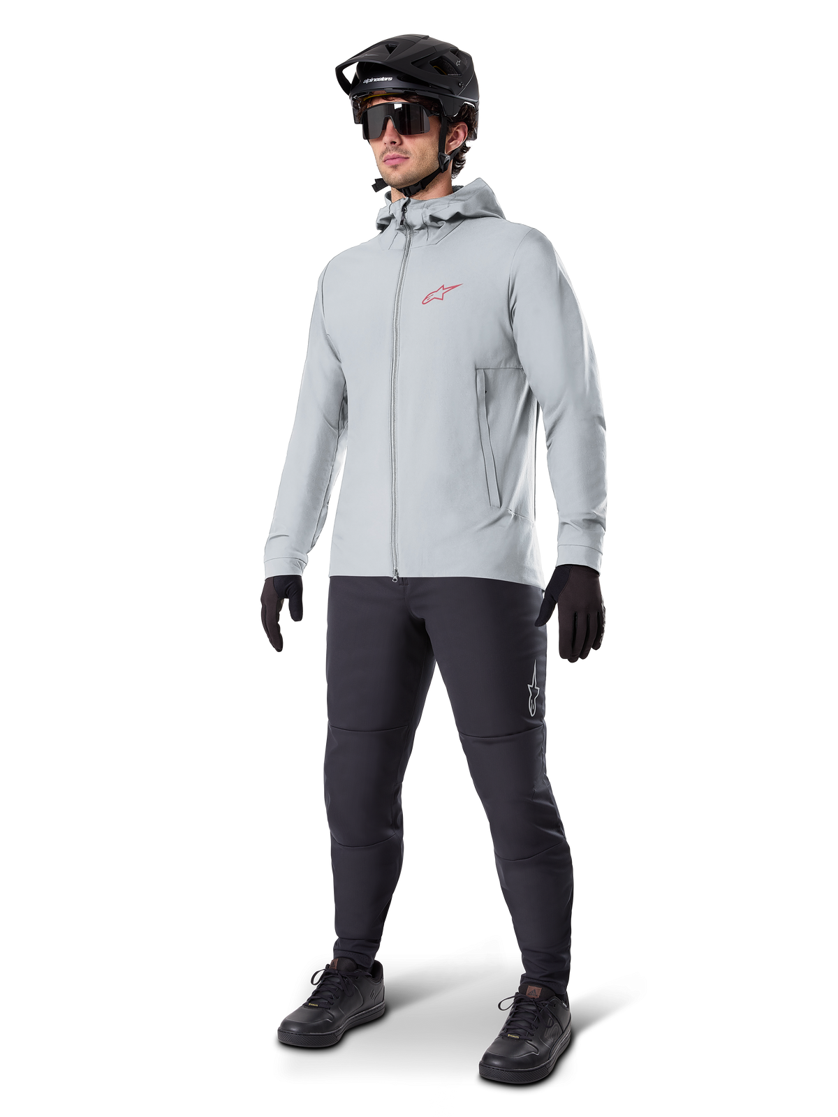 Alpinestars A-Dura Thermal Jacka i Titanium, ljusgrå cykeljacka med huva och en röd Alpinestars-logotyp på bröstet, bärs av en man med svart MTB-hjälm, solglasögon, mörkgrå byxor och svarta handskar, professionell mountainbike-utrustning.
