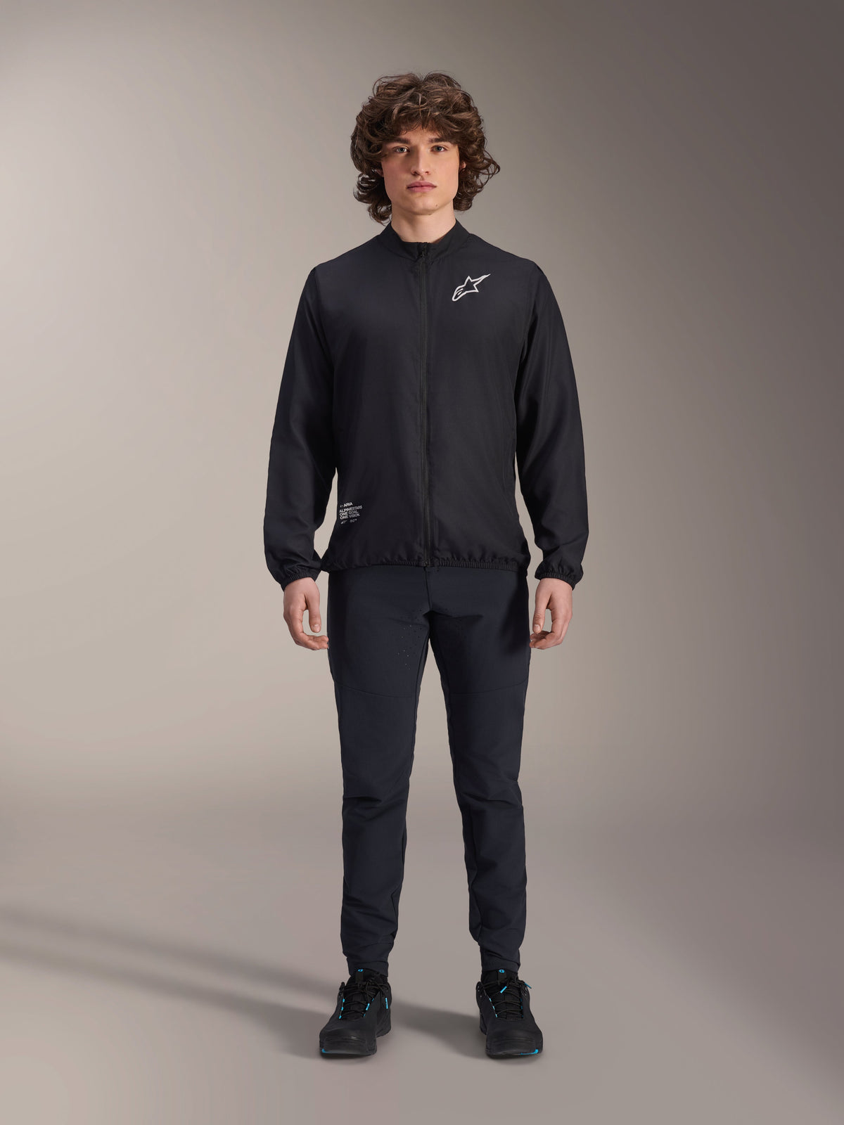 Modell iklädd Alpinestars A-Aria vindjacka i svart, med hel dragkedja fram, hög krage och vit Astars-logotyp på bröstet, matchad med svarta tekniska byxor och svarta skor, prestationsinriktade cykelkläder för mountainbike och utomhusbruk.