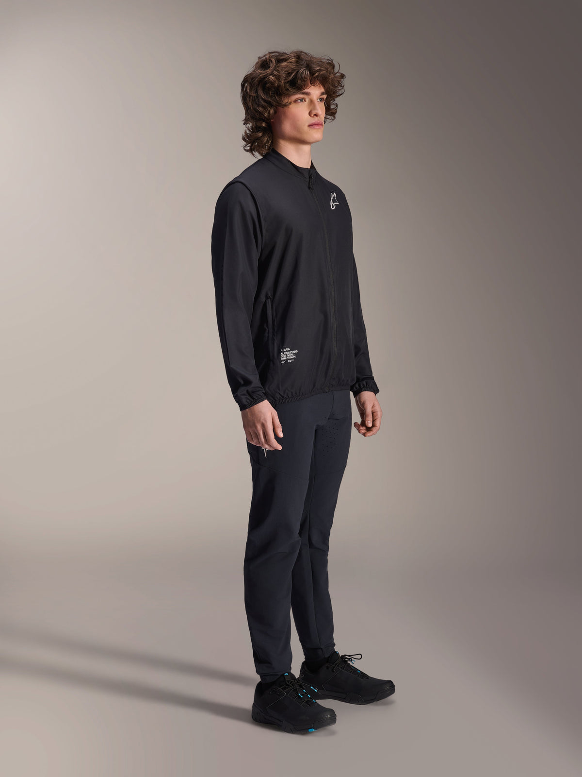 Alpinestars A-Aria Vindjacka, svart, lätt vindavvisande cykeljacka som bärs av en modell med lockigt hår, med en vit Astars-logotyp på bröstet och varumärkesdetaljer på nedre högra sidan, matchad med svarta byxor och skor, prestandapassform för mountainbike och gravelcykling