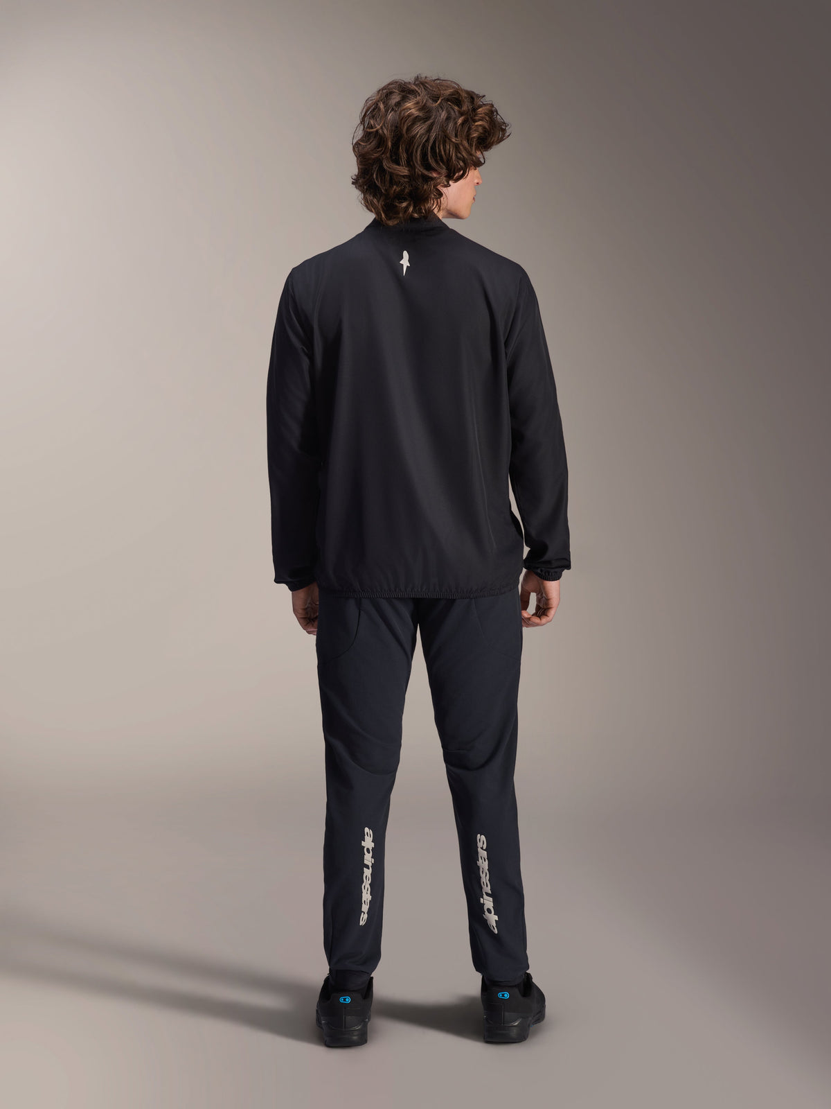 Alpinestars A-Aria vindjacka, svart, vy bakifrån, lätt vindavvisande cykeljacka med en liten vit logotyp på övre ryggen, bärs med matchande svarta byxor med vertikal Alpinestars-branding på vaderna, prestationsinriktad mountainbike-utrustning