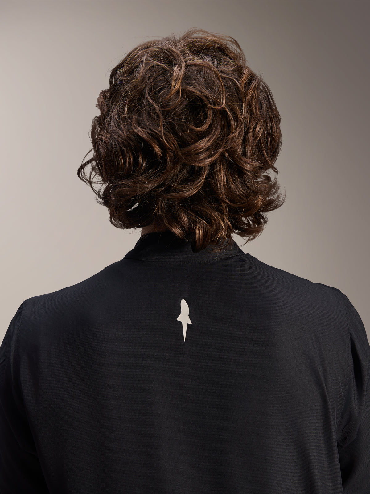 Alpinestars A-Aria vindjacka, svart, närbild bakifrån på axlar och krage, en liten vit logotyp mitt på ryggen, bärs av modell med vågigt brunt hår, minimalistisk cykelutrustning med hög prestanda