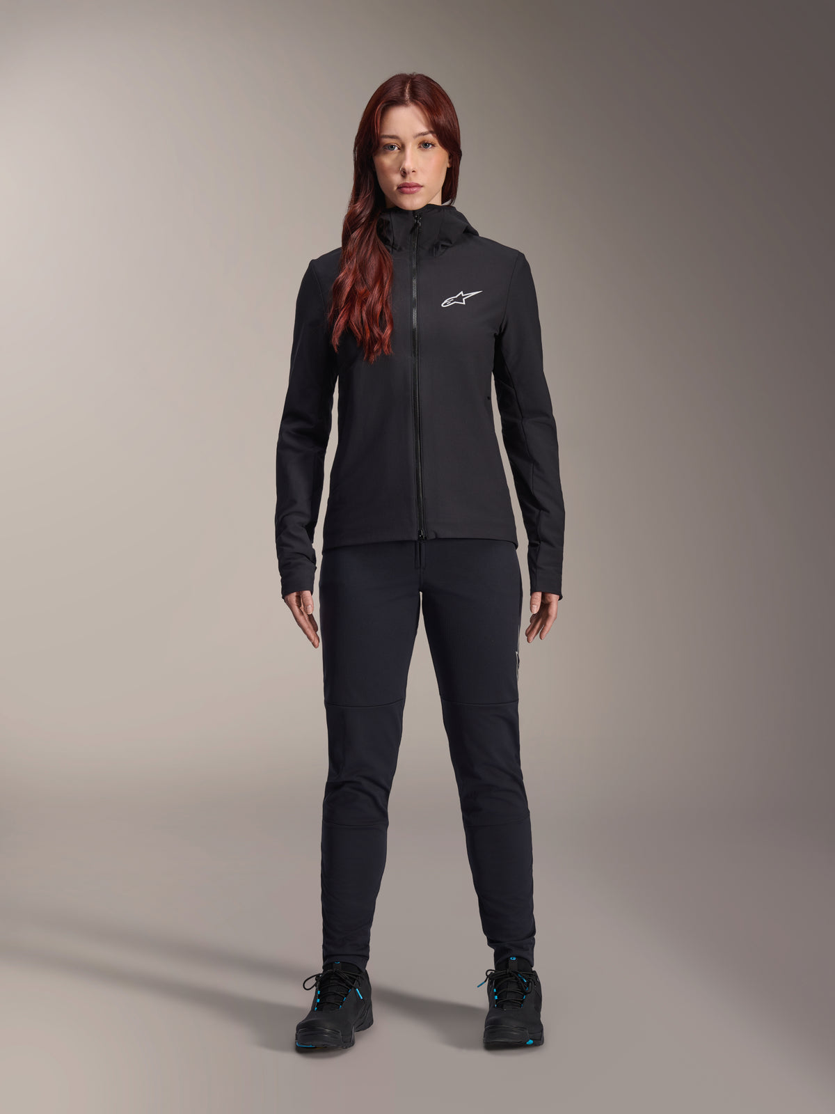 Woman wearing Alpinestars Stella A-Dura Thermal Jacket, Black, lightweight insulated cycling jacket with hood and white Astars logo on the chest, paired with black pants and athletic shoes