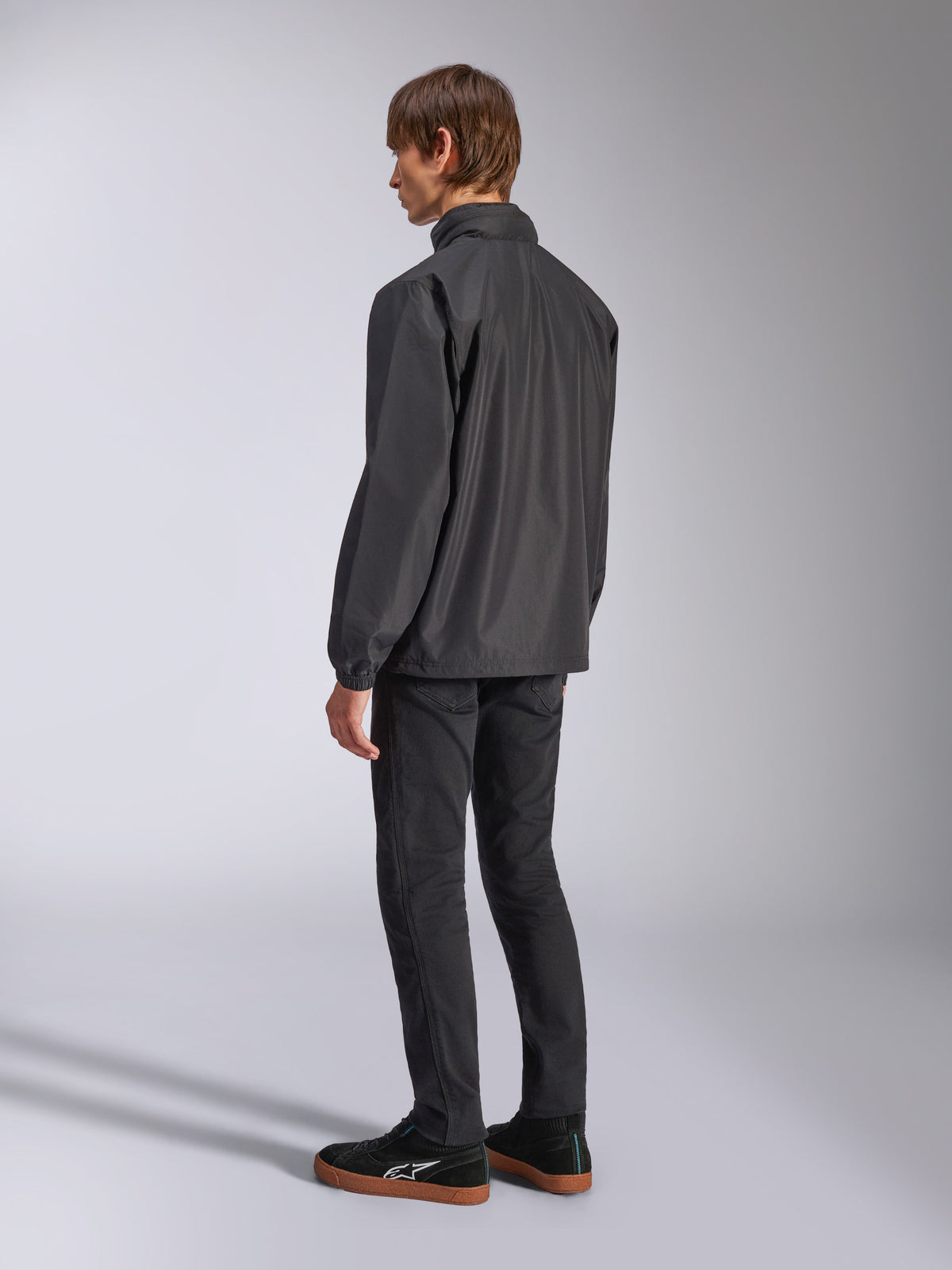 Alpinestars Overhaul Racer Jacket, Black, back three-quarter view, worn by model with black jeans and black sneakers, lightweight water-resistant rain shell jacket for casual motorsport lifestyle