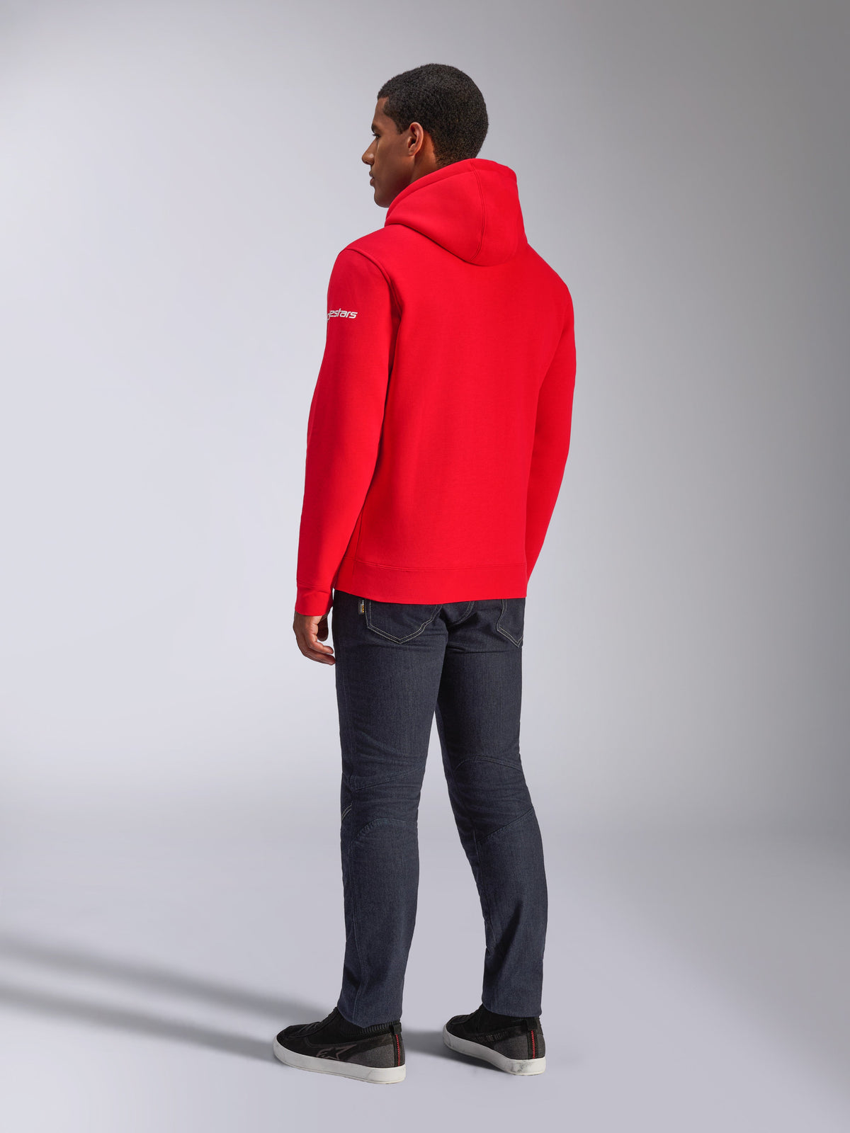 Alpinestars Frontal Hoodie, röd fleece-huvtröja i pullover-modell, vy bakifrån som visar vit wordmark-logotyp på ärmen, bärs med mörka jeans och svarta skor, avslappnad motorsport-streetwear