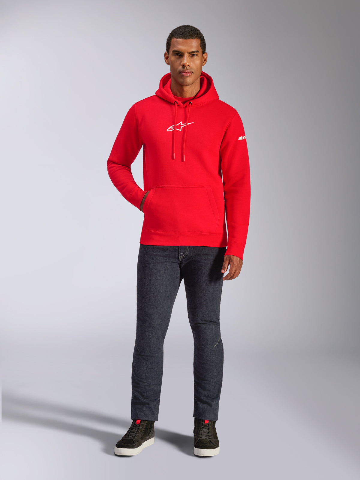 Manlig modell som bär Alpinestars Frontal Hoodie i rött, fleecehuvtröja i pullover-modell med vit Astars-logotyp på bröstet, känguruficka, huva med dragsko, kombinerat med mörkgrå jeans och svarta höga sneakers, avslappnat motorsport-streetwear