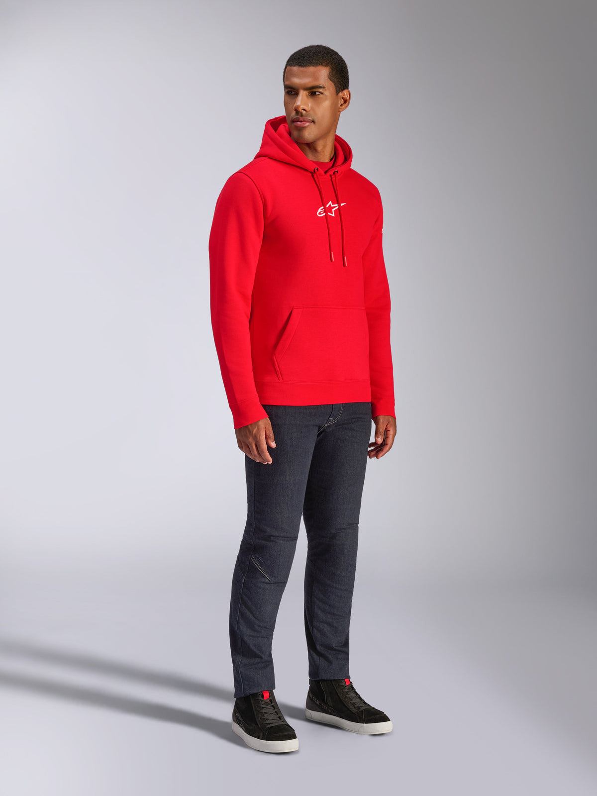 Alpinestars Frontal Hoodie, röd, manlig modell bär en fleecehuvtröja i pullover-modell med vit A-star-logotyp på bröstet, känguruficka, huva med dragsko, matchad med mörka jeans, avslappnad motorsportinspirerad streetwear