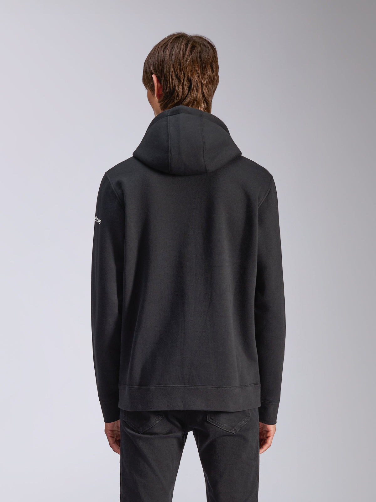 Alpinestars Frontal Hoodie, svart, baksida av en vardaglig pullover-huvtröja med självfodrad huva, ribbstickad fåll och vit wordmark-logotyp på ärmen, motorsportinspirerat streetwear