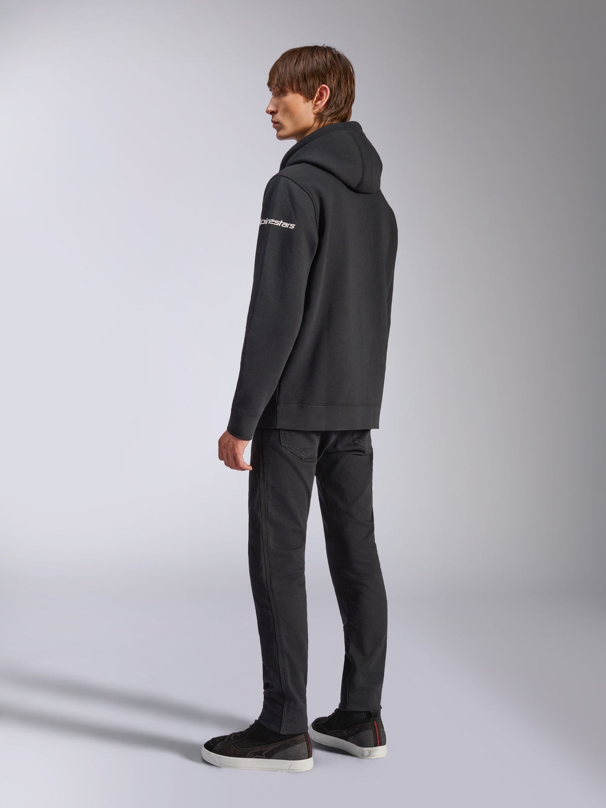 Alpinestars Frontal Hoodie, vardaglig streetwear, svart, sidovy bakifrån som visar vit wordmark-logotyp på ärmen, bärs med svarta byxor och sneakers, modern motorsportinspirerad design