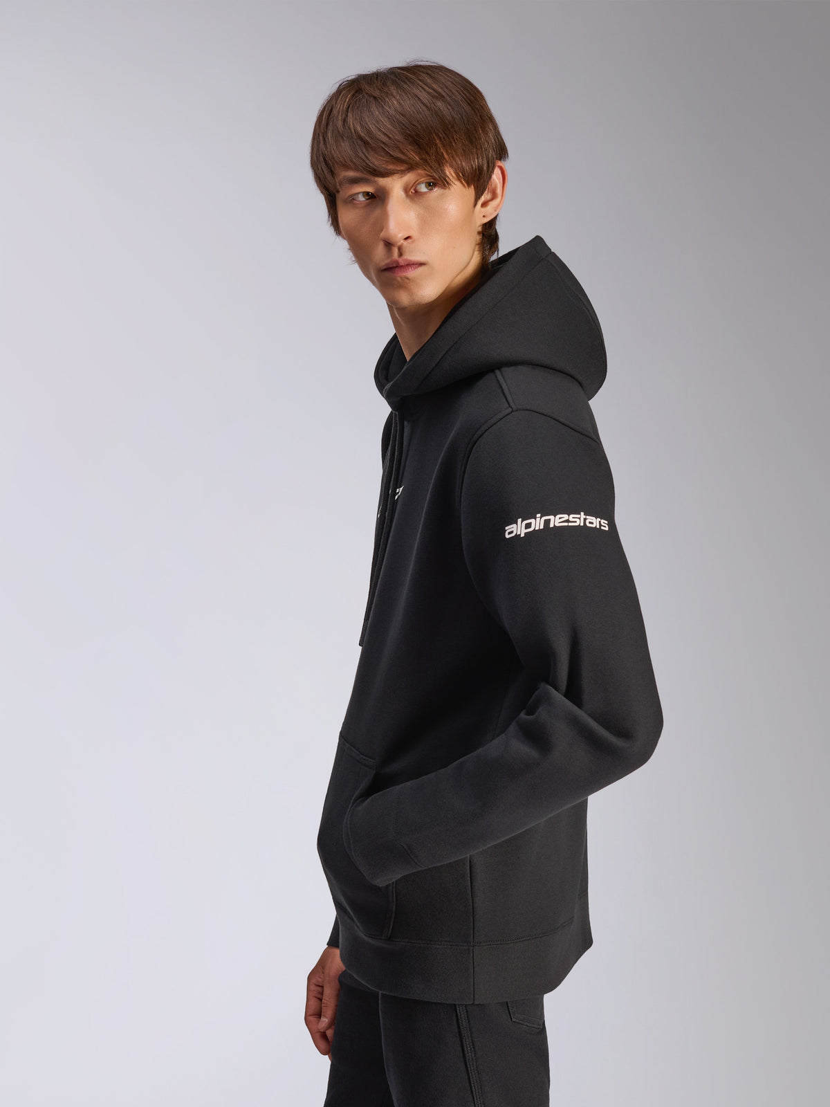 Alpinestars Frontal Hoodie, svart pullover-huvtröja i fleece, modell sedd i sidoprofil, vit Alpinestars-text på ärmen, känguruficka, vardaglig motorsport-inspirerad streetwear