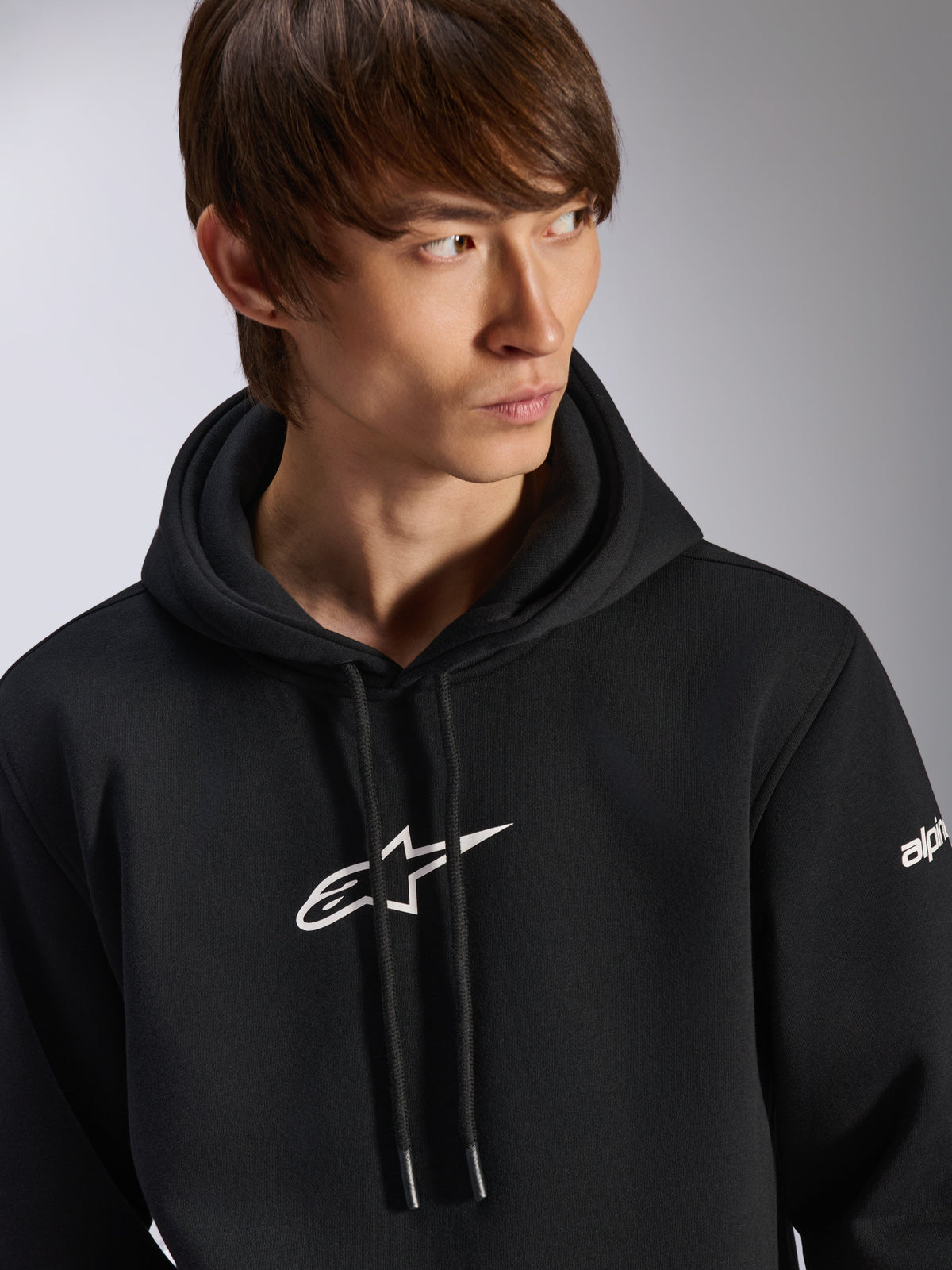 Alpinestars Frontal Hoodie, svart pullover-huvtröja i fleece, vit Astar-logotyp på bröstet och wordmark-logotyp på ärmen, huva med dragsko, bärs av en manlig modell, casual motorsport-streetwear