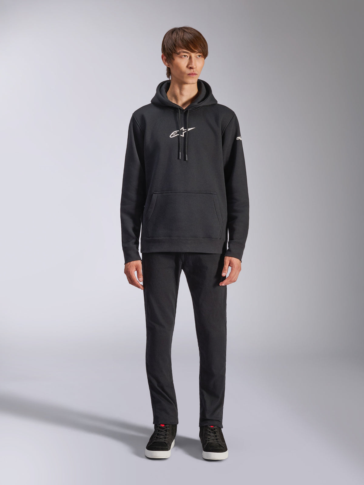 Alpinestars Frontal Hoodie, svart pullover-huvtröja i fleece, vit Alpinestars-logotyp på bröstet och ärmen, känguruficka, huva med dragsko, bärs med svarta byxor, avslappnad motorsportinspirerad design