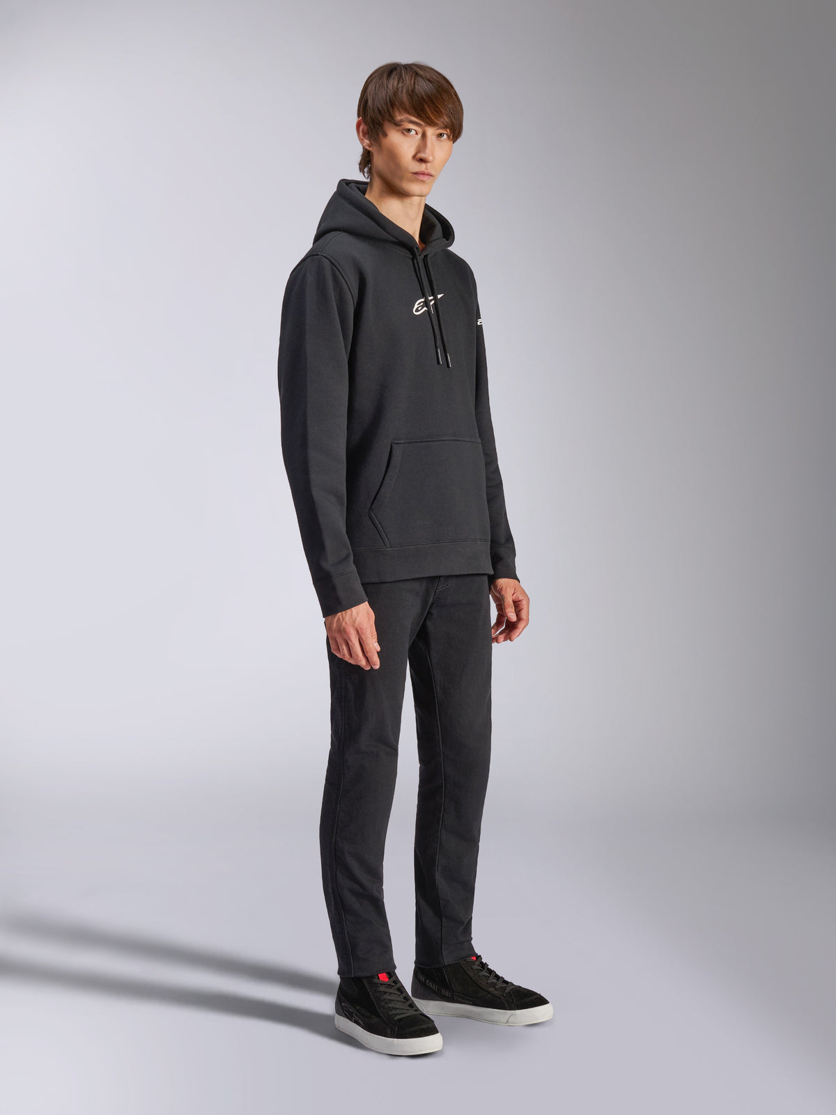 Alpinestars Frontal Huvtröja, svart, pullover fleece-huvtröja med vit HD-logotyp på bröstet och wordmark-logotyper på ärmarna, känguruficka, stylad med svarta byxor och sneakers, avslappnad motorsportinspirerad streetwear