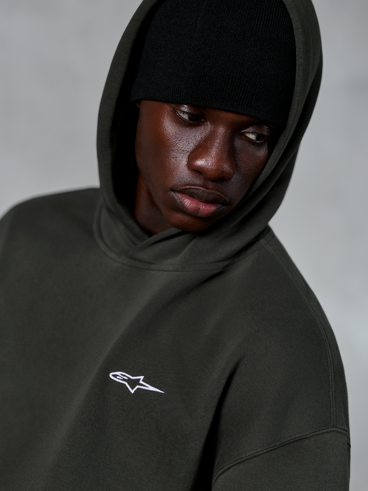 Alpinestars Essentials Ageless huvtröja, Off Black, närbild på modell med en svart mössa under huvan, med en liten vit stjärnlogotyp på bröstet, avslappnad motorsportinspirerad streetwear