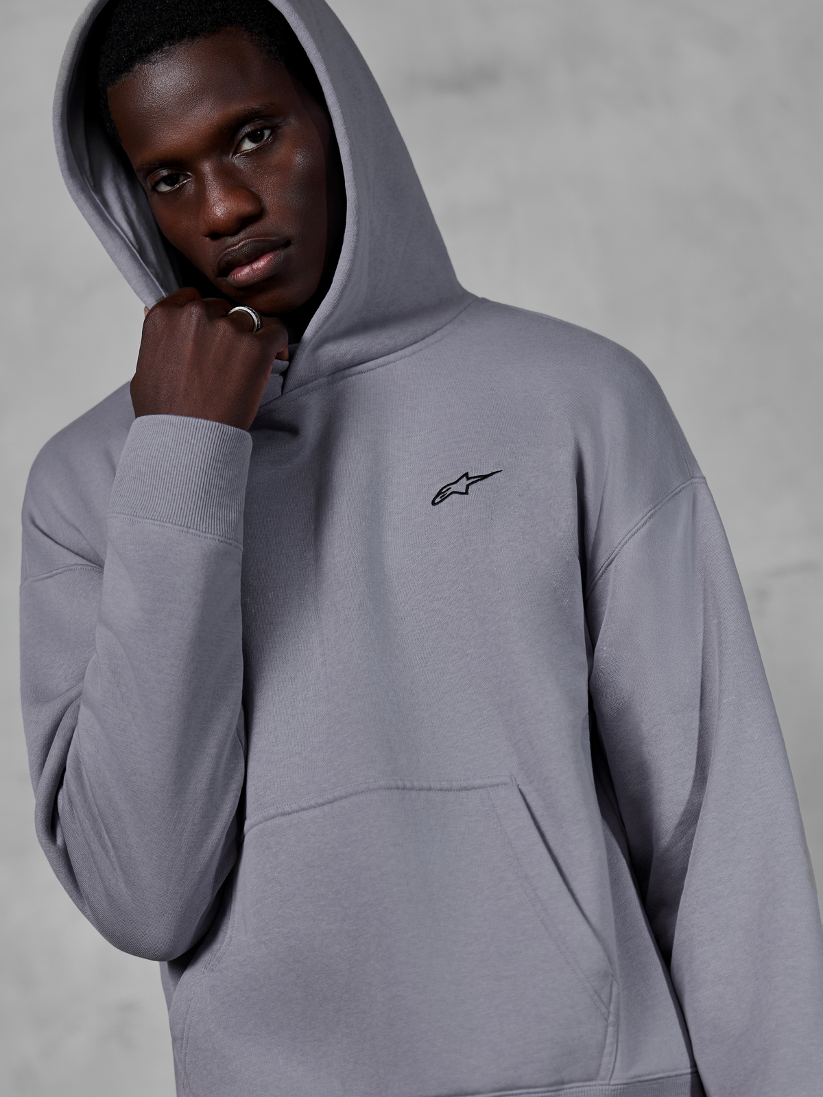 Alpinestars Essentials Ageless Hoodie, Silver, manlig modell som bär huvtröjan med huvan uppfälld, liten svart Alpinestars-logotyp på bröstet, känguruficka, ribbade muddar, avslappnad lifestyle streetwear