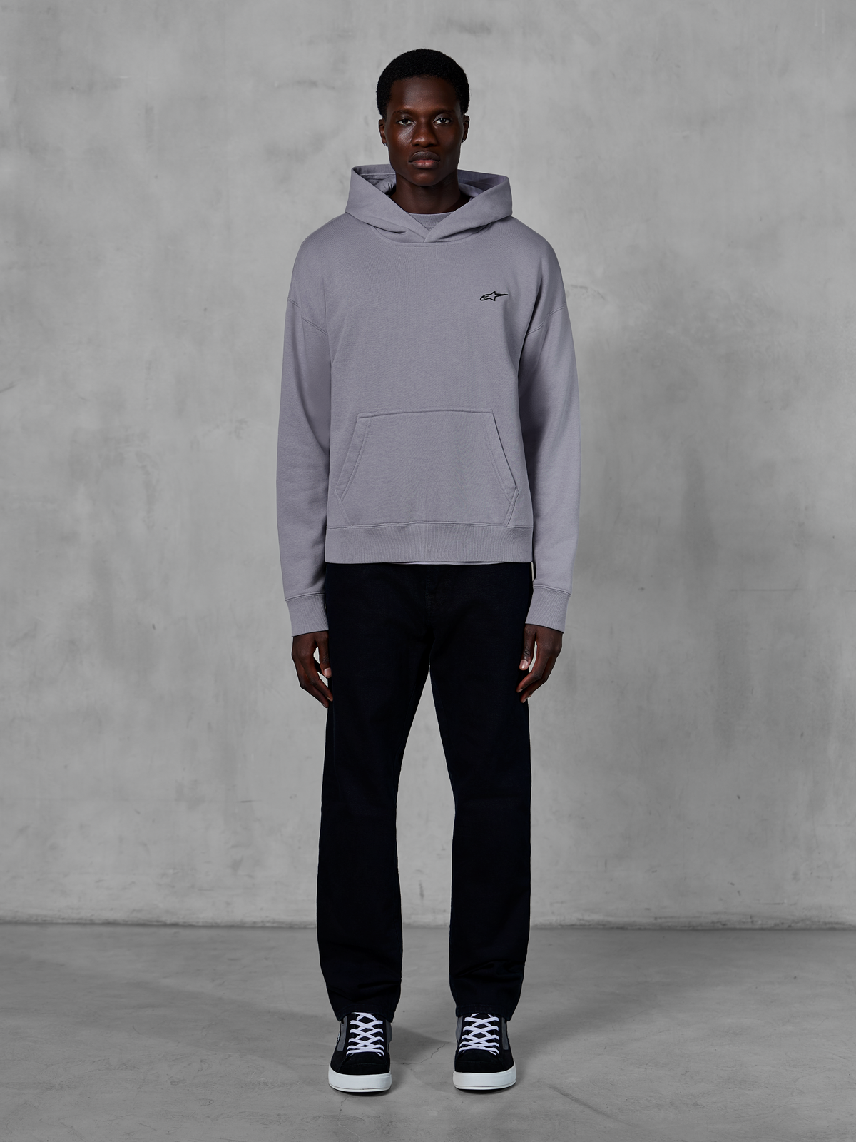 Alpinestars Essentials Ageless Hoodie, silver, manlig modell i avslappnad streetwear, liten svart Alpinestars-logotyp på bröstet, känguruficka framtill, minimalistisk design, matchad med svarta byxor och sneakers.