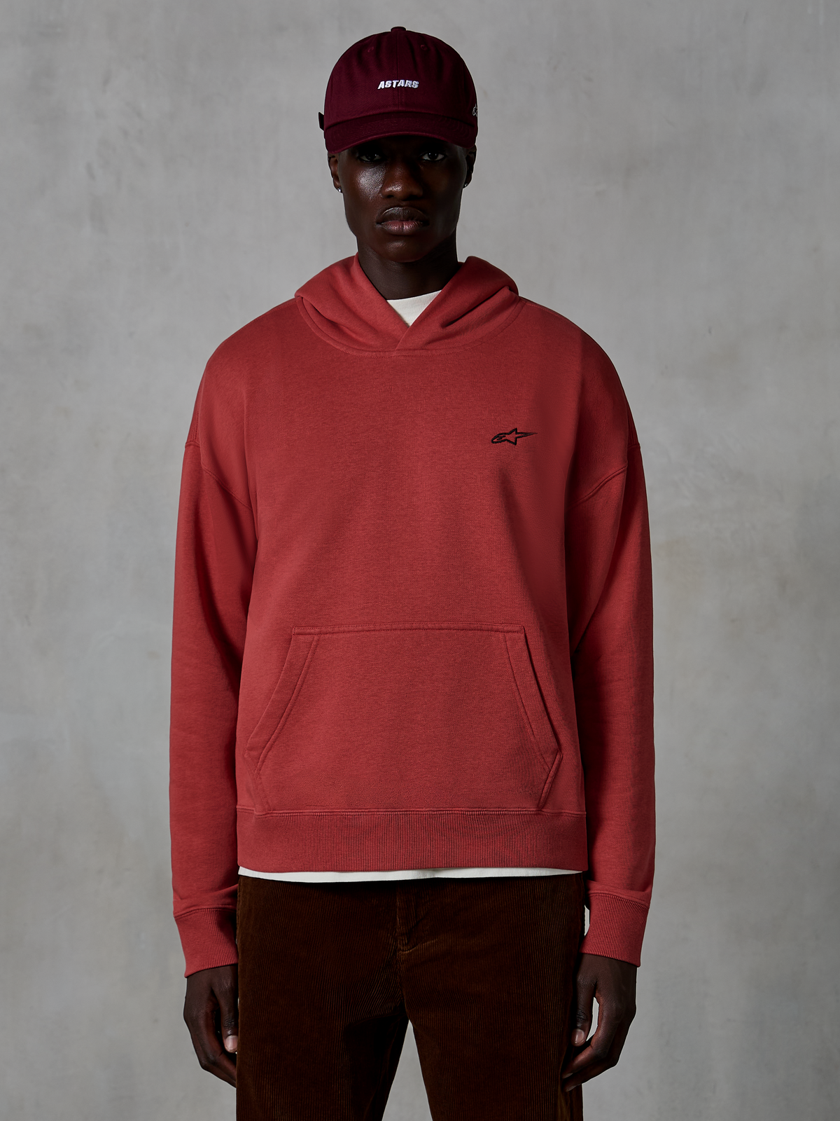 Alpinestars Essentials Ageless Hoodie, casual streetwear, Brick red color, featuring a small black star logo on the chest, kangaroo pocket, stone-wash finish, worn by a model with a maroon cap and brown corduroy pants.