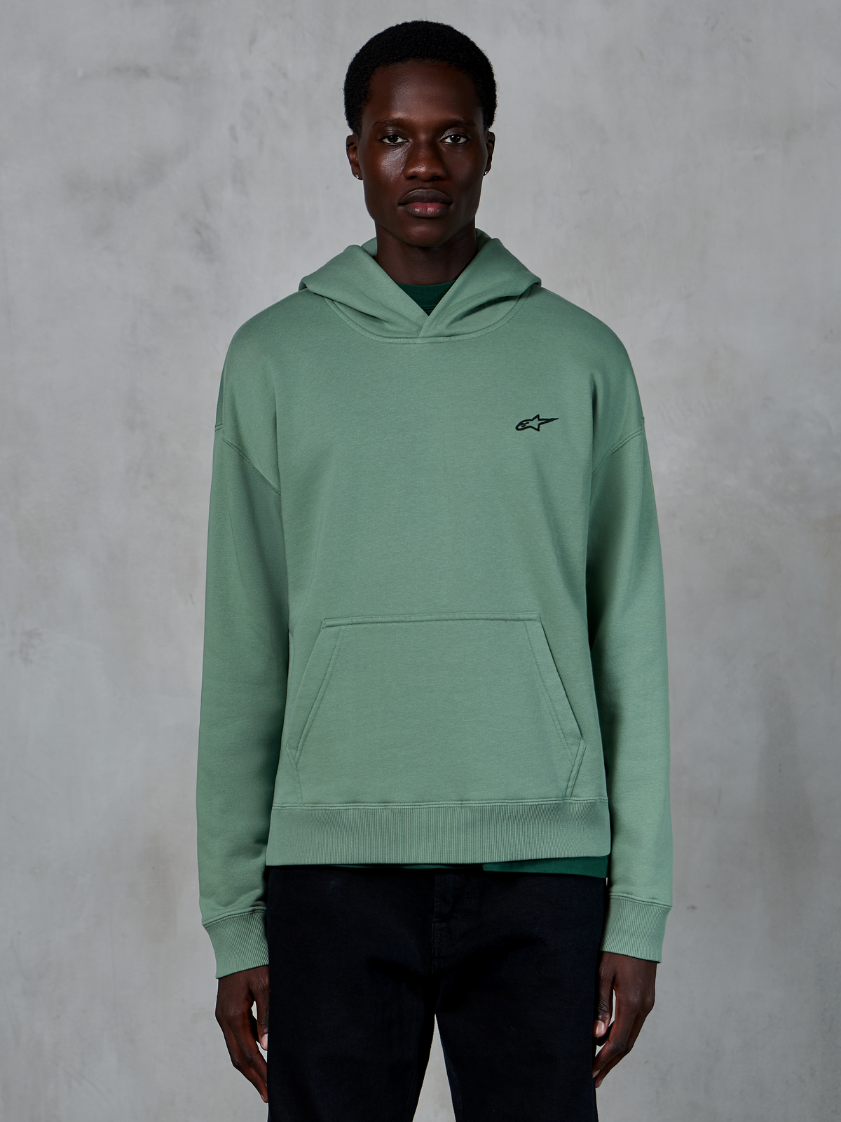 Alpinestars Essentials Ageless Hoodie, cactusgrön, ledig fleecehuvtröja med stentvättad finish, har en liten svart stjärnlogotyp på bröstet och en känguruficka fram, bärs av en modell i en ren livsstilsmiljö