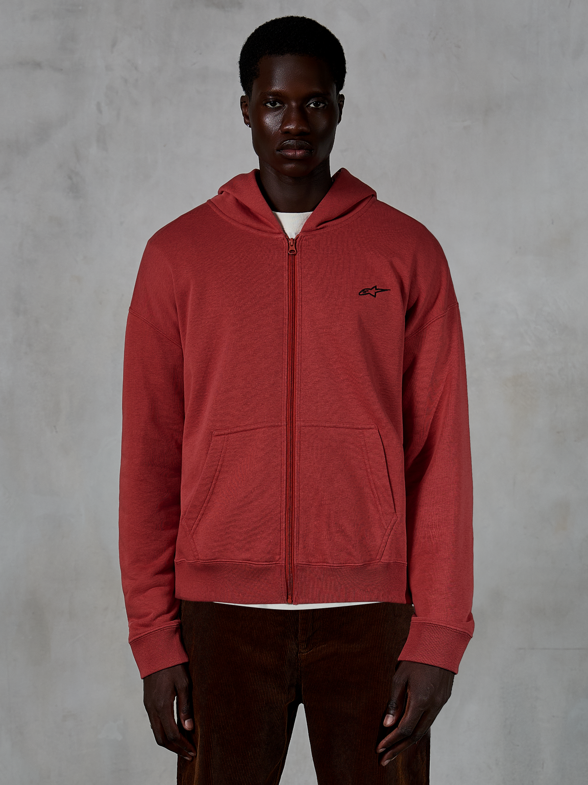 Alpinestars Essentials Ageless Zip Hoodie, tegelröd luvtröja med dragkedja, bärs av modell med bruna manchesterbyxor, liten svart logotyp på bröstet, avslappnad passform, vardaglig streetwear