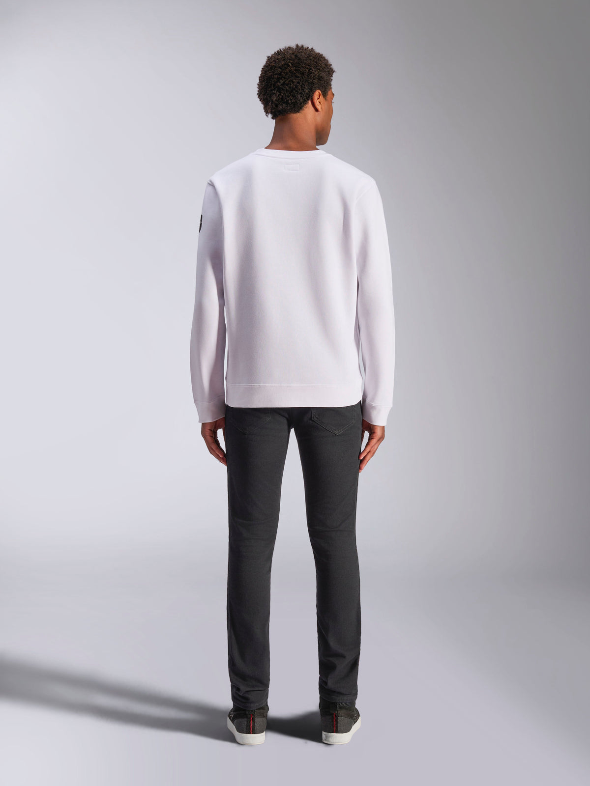 Alpinestars Bettern Crew, white crewneck sweatshirt, back view on model, paired with dark jeans and sneakers, casual motorsport-inspired streetwear