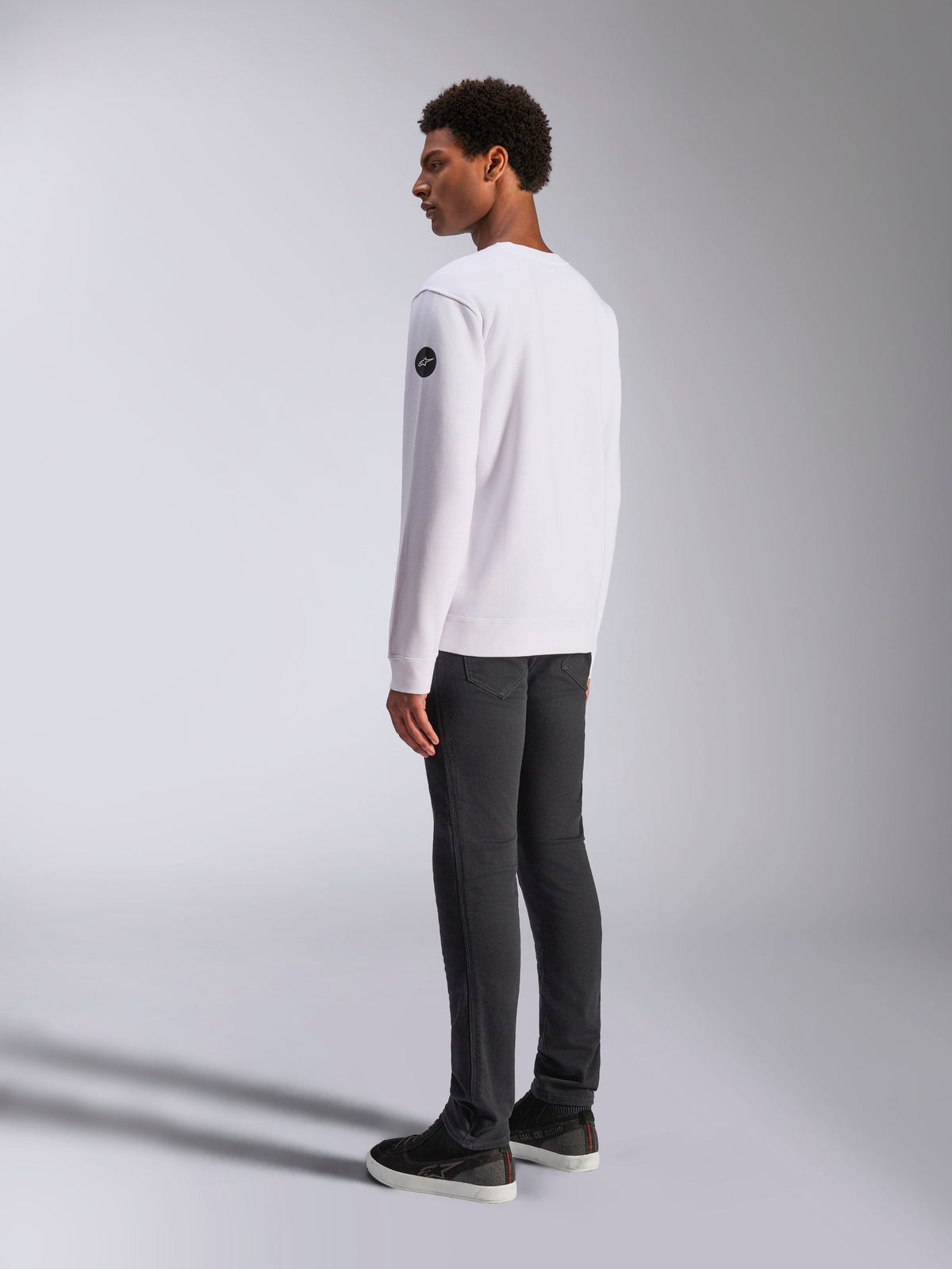 Alpinestars Bettern Crew, White Crewneck Sweatshirt, back and side view, black circular logo on the sleeve, worn with dark grey pants and black sneakers, casual motorsport lifestyle streetwear