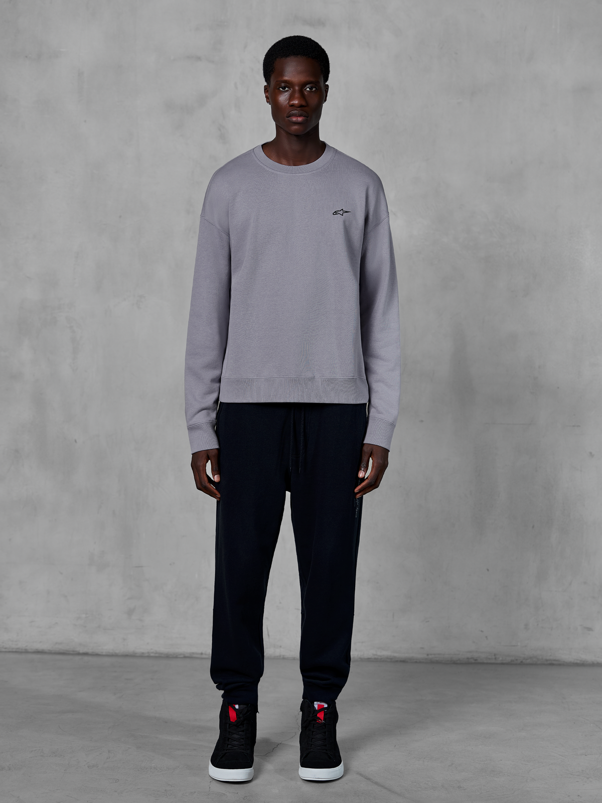 Alpinestars Essentials Ageless Crew, silverfärgad crewneck-tröja, bärs av en modell med svarta mjukisbyxor och svarta sneakers, med en liten svart Alpinestars-logotyp på bröstet, avslappnat motorsportinspirerat streetwear