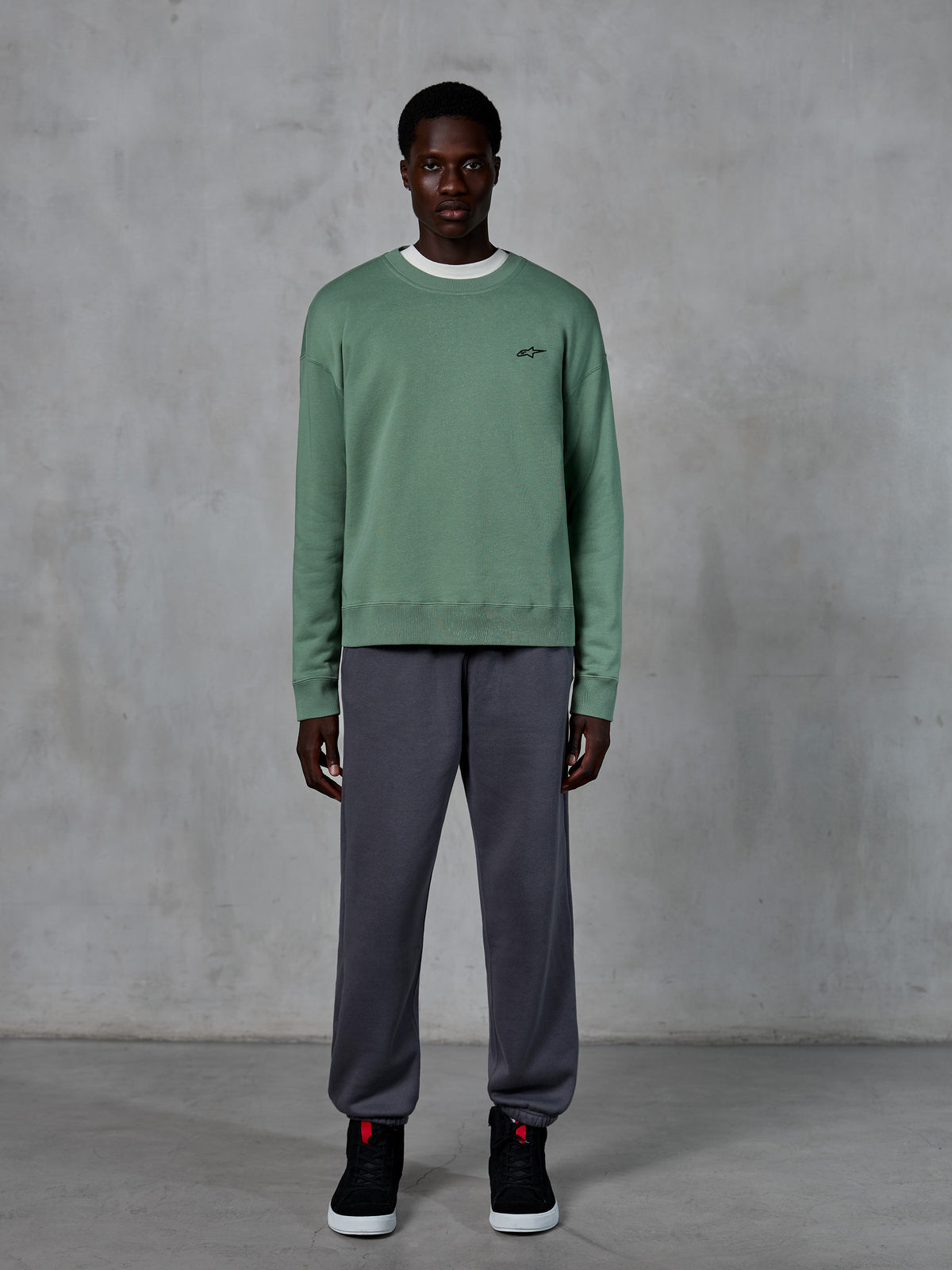 Alpinestars Essentials Ageless Crew, crewneck-tröja i Cactus-grön med en liten svart logotyp på bröstet, bärs av en modell med grå mjukisbyxor och svarta sneakers, avslappnad motorsportinspirerad streetwear