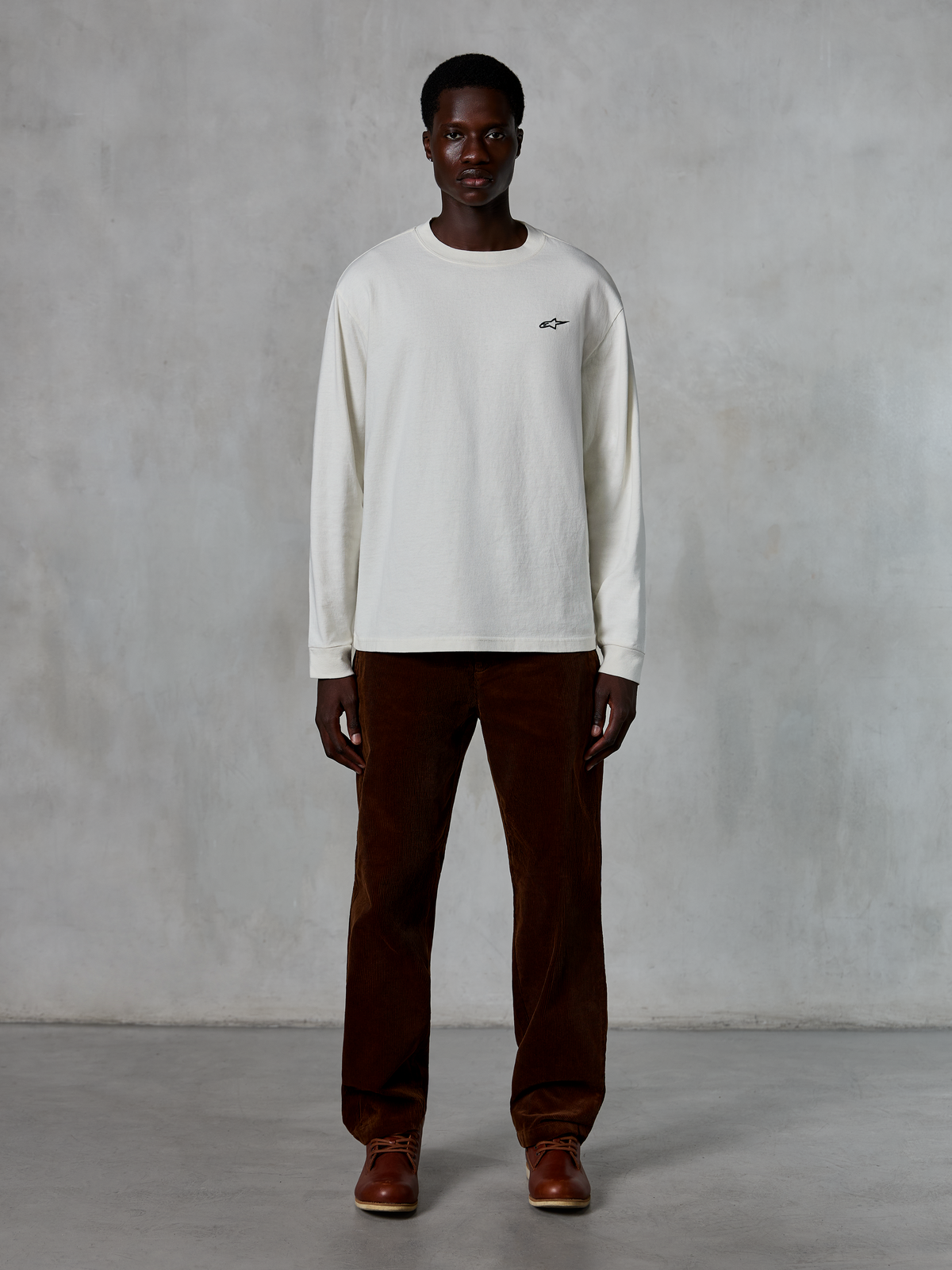 Alpinestars Essentials Ageless LS Tee, långärmad t-shirt, Off-White, bärs av modell med bruna manchesterbyxor och bruna läderkängor, med en liten svart logotyp på bröstet, avslappnad modern passform, casual lifestyle streetwear
