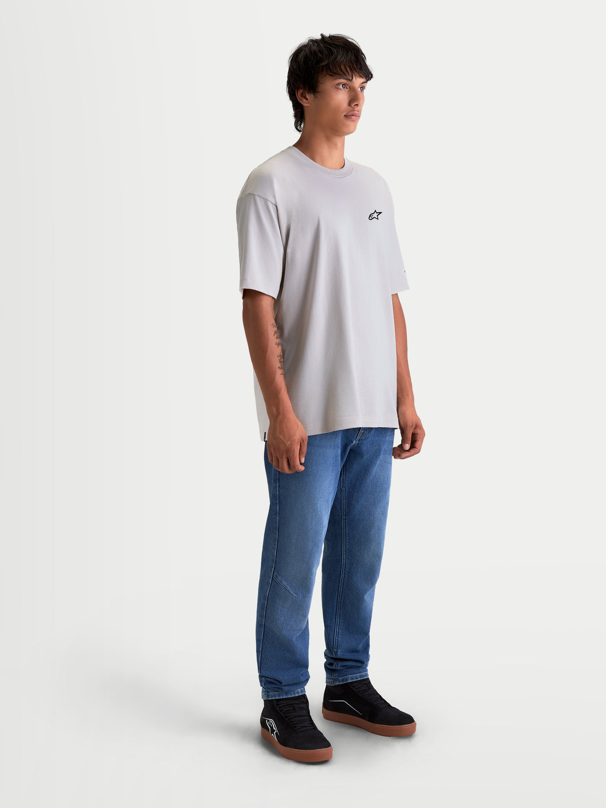 Alpinestars Mandate Oversized CSF t-shirt, cementgrå kortärmad sportswear t-shirt med avslappnad oversized passform och en liten svart logotyp på bröstet. Tillverkad i en mjuk bomulls-polyesterblandning för vardagskomfort. Visas på en manlig modell med blå jeans och svarta sneakers.