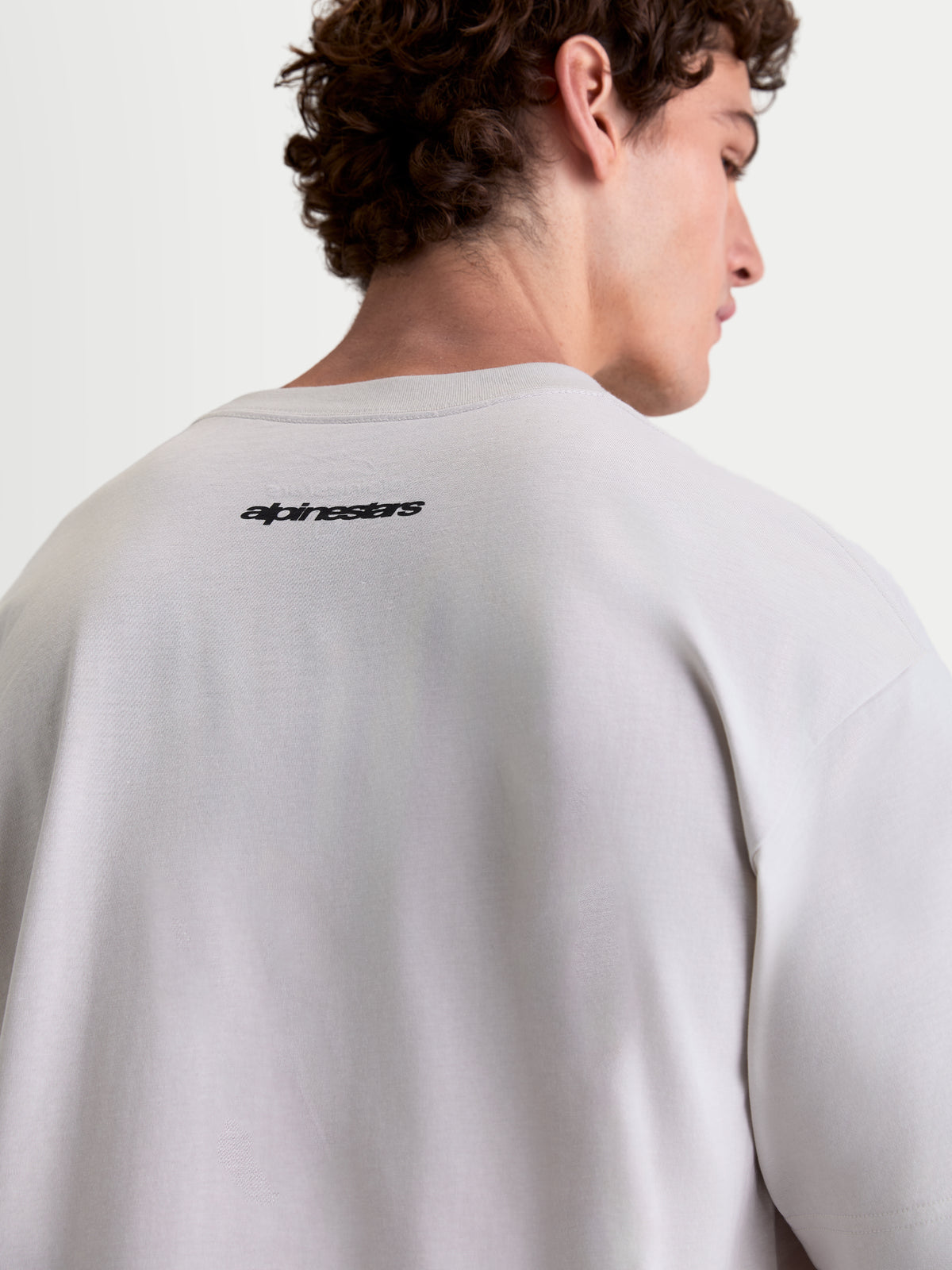 Alpinestars Prelude Oversized CSF t-shirt, kortärmad sportswear-t-shirt, ryggvy på modell som visar Alpinestars ordmärkesgrafik, cementgrå och svart, avslappnad oversized passform i bomull- och polyesterblandning.