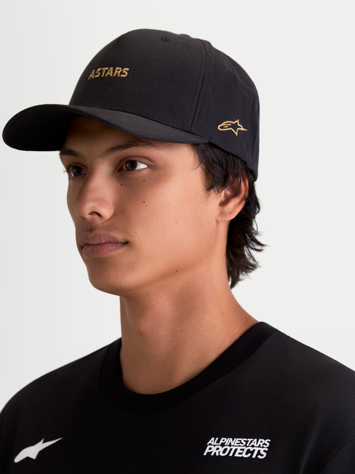 Alpinestars County Canvas Hatt buren av modell, svart sportswearkeps med guld-ASTARS-logotyp och sidobroderi, böjd skärm och strukturerad krona, oxfordväv, motorsportinspirerat vardagsplagg.