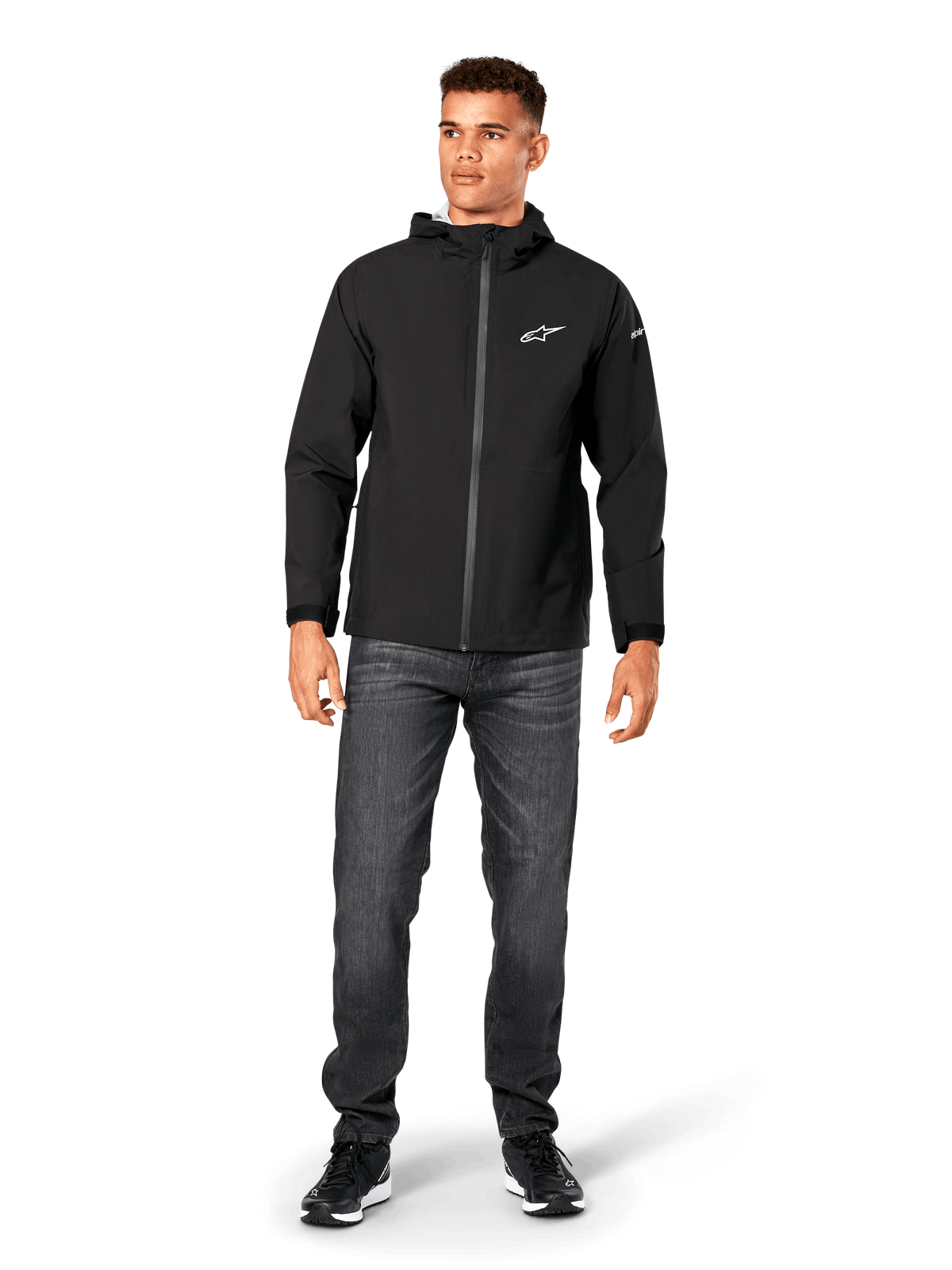 Alpinestars Kascade Rain Jacket, Men's Rain Shell, Black, worn with dark grey jeans and black sneakers, featuring a hooded design, coated front zipper, and white Alpinestars logo on the chest, casual motorsport-inspired streetwear