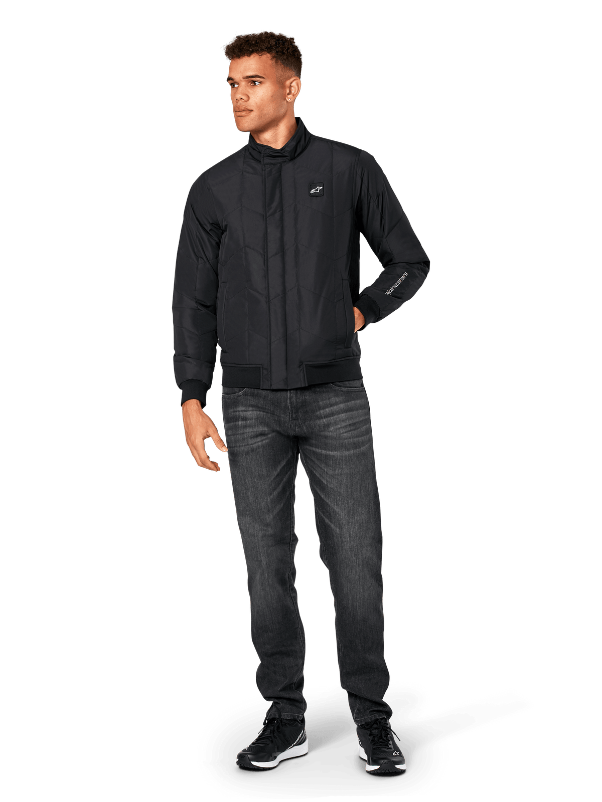 Alpinestars Precedent Jacket, quiltad sportjacka, svart, bärs med mörkgrå jeans och svarta sneakers, med ett vinklat quiltmönster, låg ståkrage, ribbstickade muddar och nederkant, vitt logomärke på bröstet och broderi på ärmen.