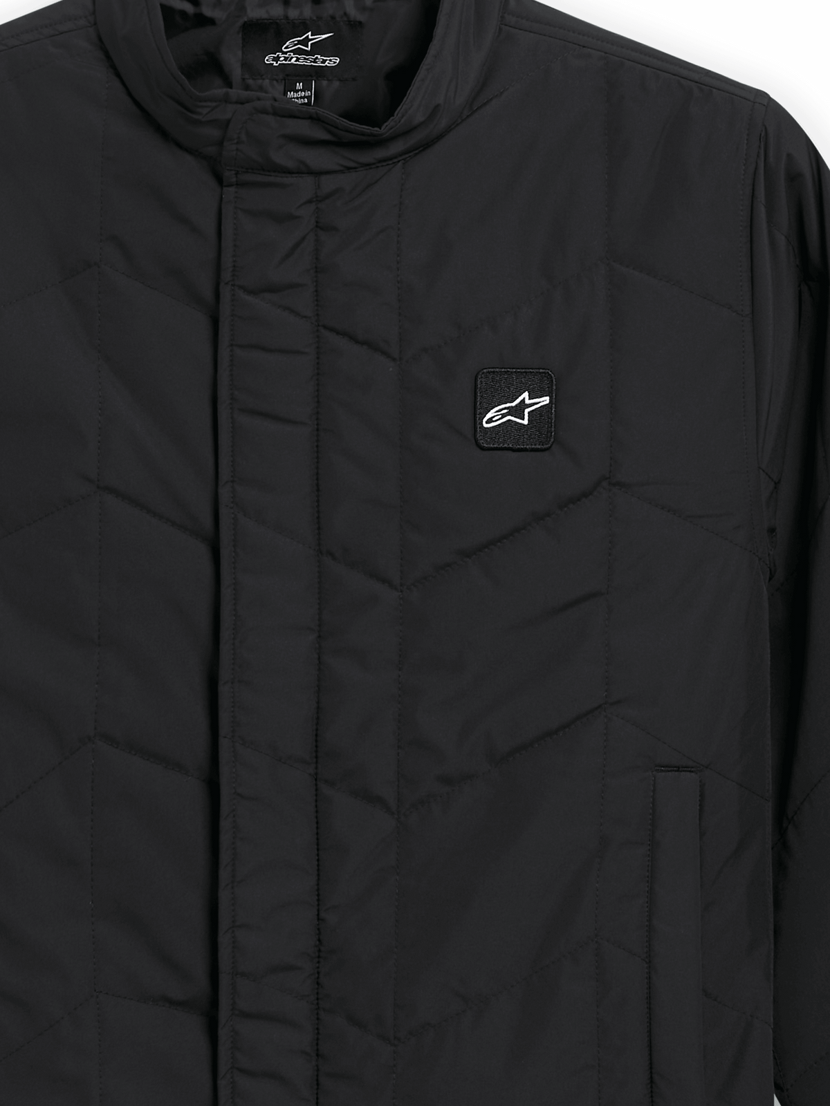 Alpinestars Precedent Jacka, quiltad sportswearjacka, svart, närbild som visar ett ikoniskt vinklat quiltmönster, låg ståkrage, täckt dragkedja framtill och ett litet logomärke på bröstet