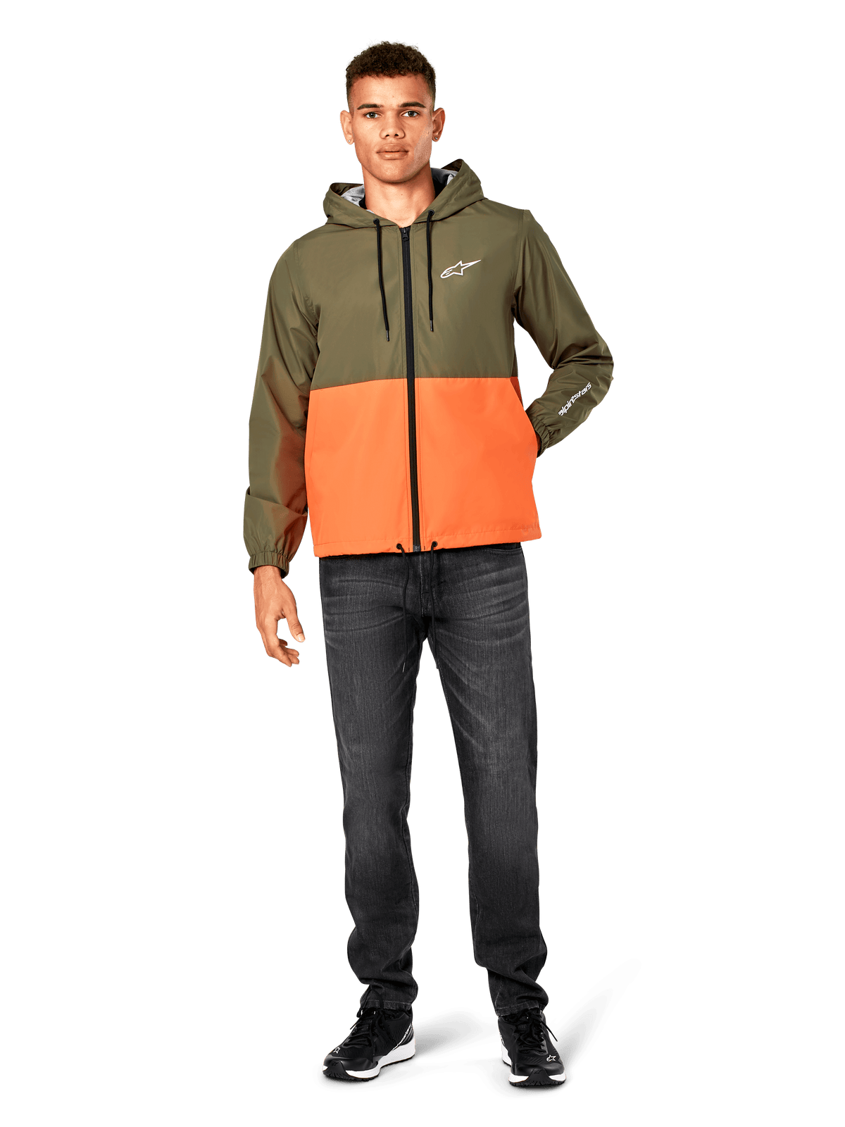 Alpinestars Speeder Windbreaker Jacket, blockfärgad i militärgrönt och orange, lätt jacka med huva, svarta dragsnören och dragkedja framtill, vit logotyp på bröstet och ärmen, bärs med mörka jeans och svarta sneakers, avslappnad sportstil.