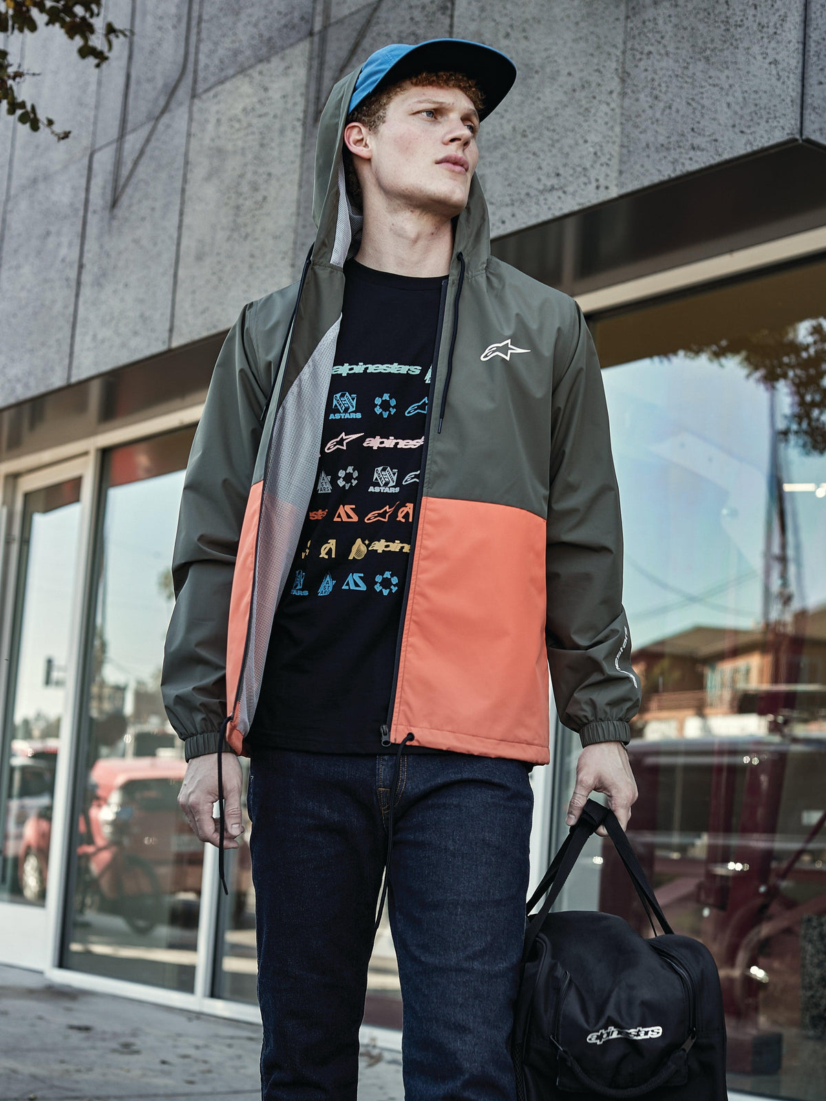 Man som bär Alpinestars Speeder Windbreaker Jacket i Military Orange, militärgrön och orange blockfärgad design, huva med meshfoder, matchad med en svart t-shirt med grafisk logotyp, blå jeans och en svart Alpinestars duffelväska, sportig livsstilslook