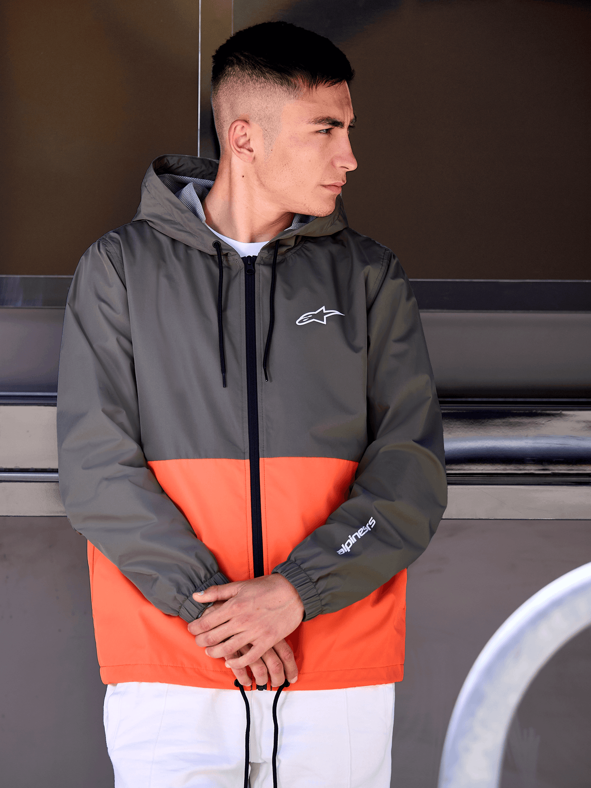 Man som bär Alpinestars Speeder vindjacka, blockfärgad design i militärorange och mörkgrönt, ytterplagg med huva och vita logotyper på bröst och ärm, kombinerat med vita byxor för en motorsportinspirerad livsstilslook