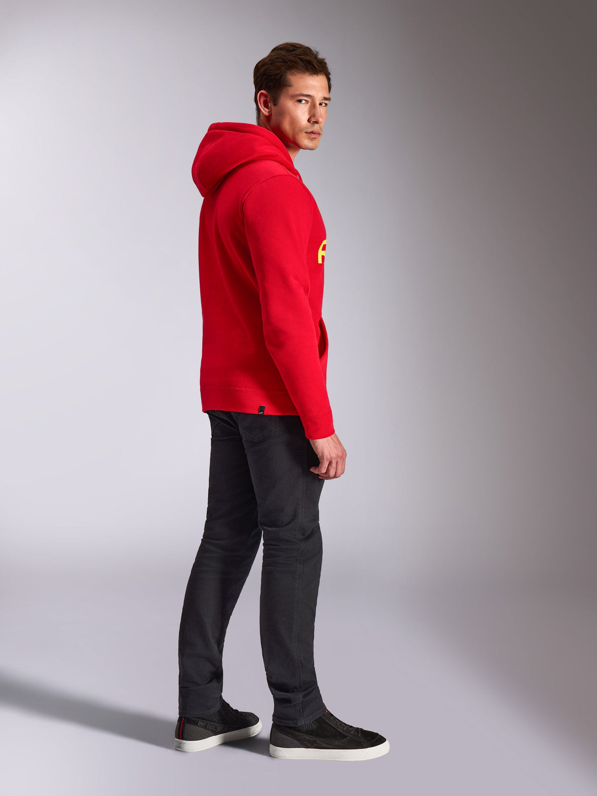 Alpinestars Linear Wordmark V3 Hoodie, röd, avslappnat streetwear, modell sedd från sidan och bakifrån bärandes en röd huvtröja med gul logotyp, mörkgrå jeans och svarta sneakers, studio-livsstilsbild