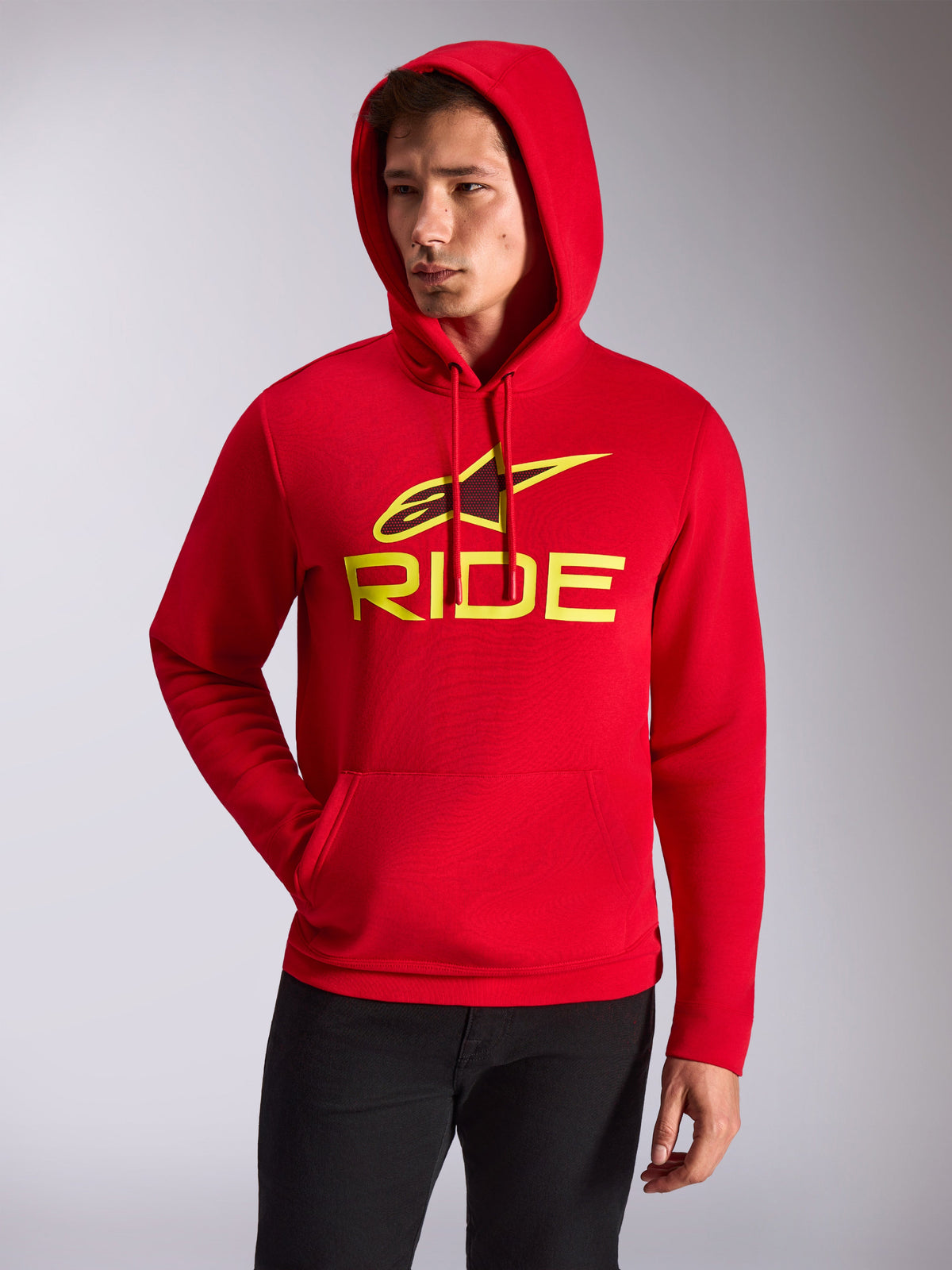 Alpinestars Ride V3 Hoodie, manlig modell i röd fleecehuvtröja, neongul RIDE-logotyp och Alpinestars-stjärna på bröstet, vardaglig motorsport-streetwear, känguruficka fram, bärs med svarta byxor.