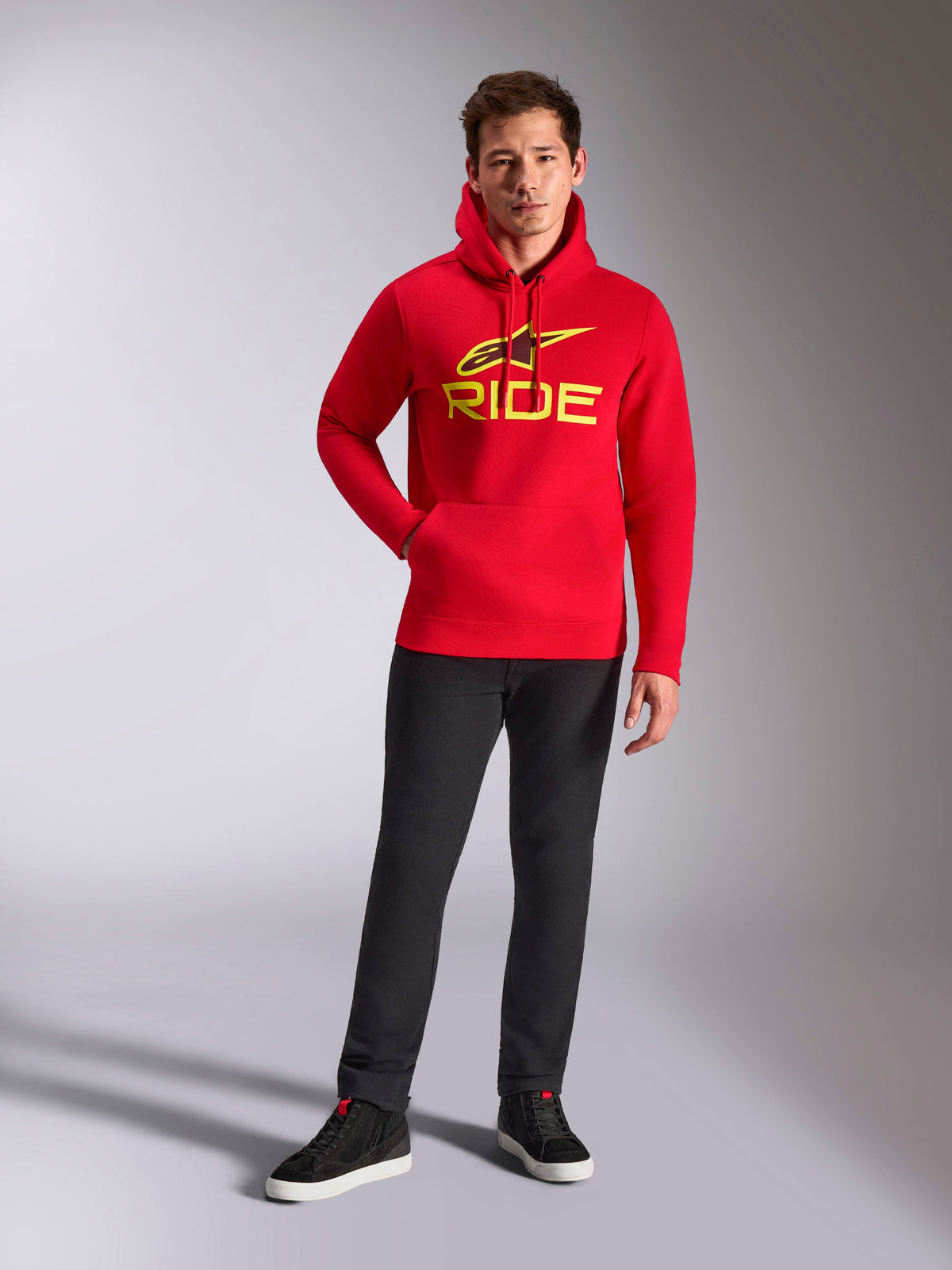 Alpinestars Ride V3 huvtröja, röd pullover i fleece, lysande gul RIDE-logotyp med Astars-symbol på bröstet, bärs med svarta byxor och svarta höga sneakers, avslappnad motorsport-inspirerad streetwear