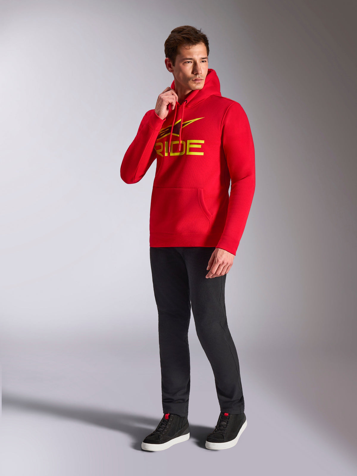 Alpinestars Ride V3 Hoodie, röd huvtröja, gul och svart 'RIDE'-logotyp på bröstet, bärs av modell med svarta byxor och svarta sneakers, avslappnad motorsportinspirerad streetwear