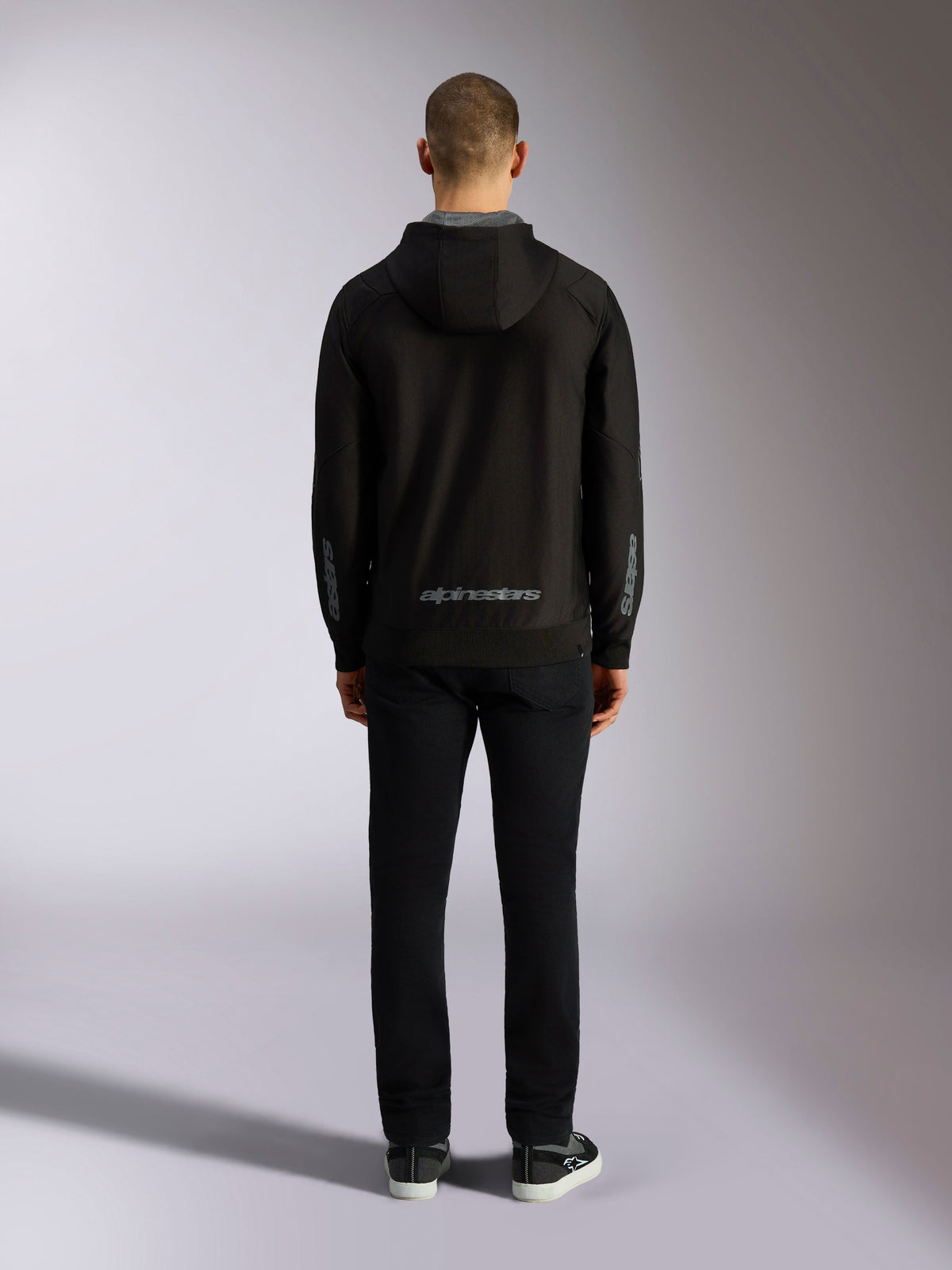 Vy bakifrån av Alpinestars Sessions V3 Hoodie i Black Grey, med ett grått Alpinestars-ordmärke på nedre ryggen och astars-logotyper på underarmarna, buren av en modell med svarta byxor, avslappnat motorsport-inspirerat streetwear