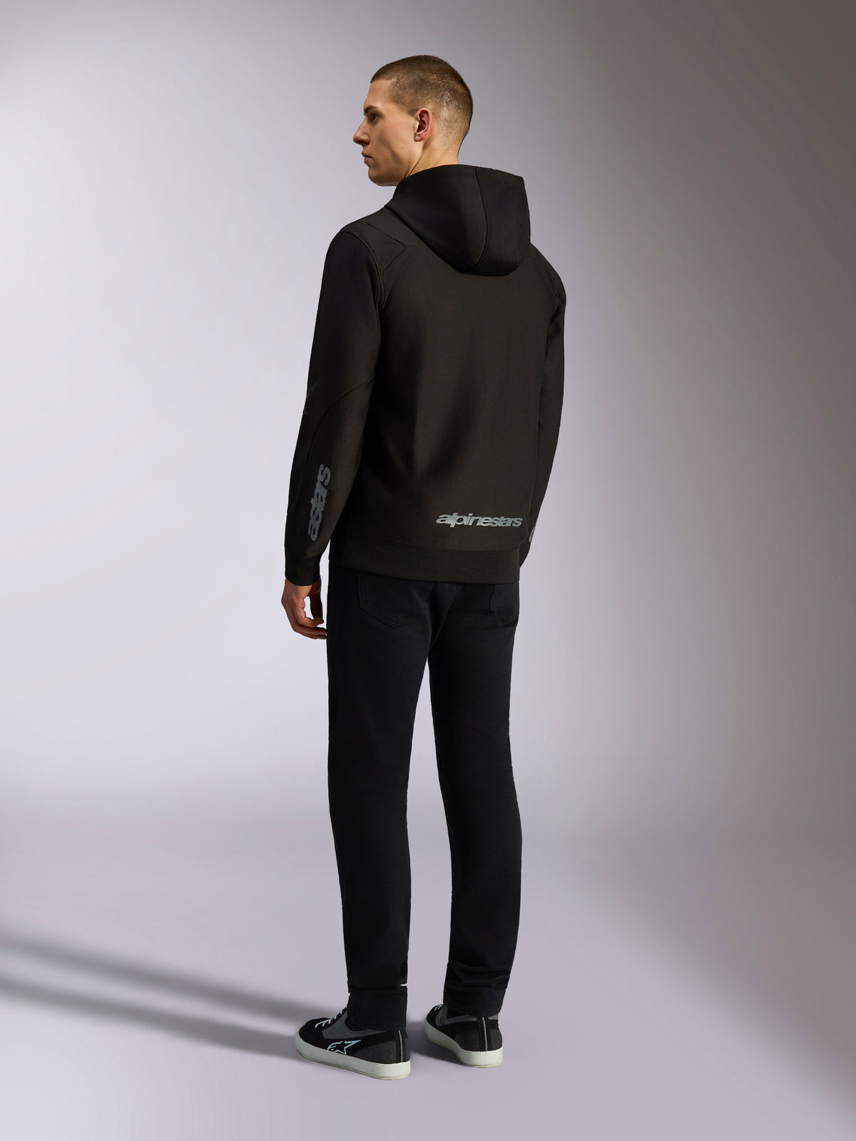 Alpinestars Sessions V3 Hoodie, svart casual streetwear, sedd bakifrån på en modell, med grå alpinestars-ordlogotyp på nedre ryggen och Astars-logotyp på ärmen, huvtröja med dragkedja fram och modern motorsportinspirerad design.