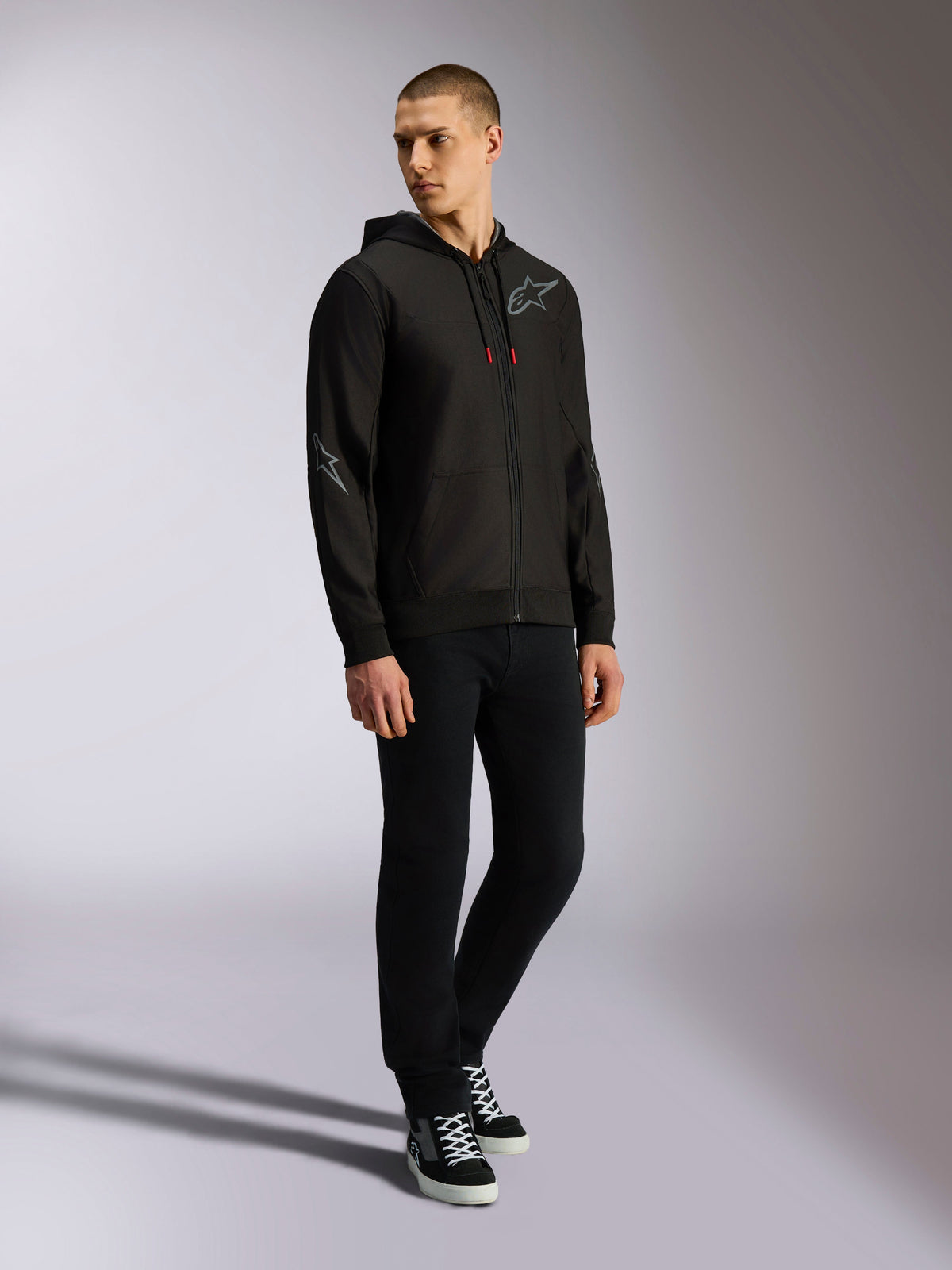 Alpinestars Sessions V3 Hoodie, huvtröja i polyfleece med dragkedja fram, svart och grå, grå logotyper på bröst och ärmar, huva med dragsnören med röda ändar, helkroppsbild av modell i avslappnad motorsport-streetwear