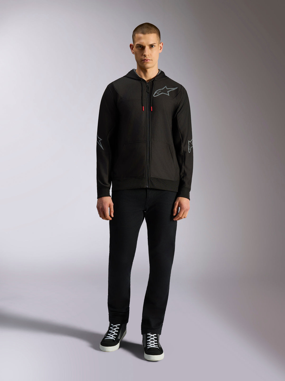 Alpinestars Sessions V3 huvtröja, svart huvtröja med dragkedja och grå Astars-logotyp på bröstet, grafik på armbågar och underarmar, röda detaljer på dragsnöret, bärs av en manlig modell med svarta byxor och sneakers, avslappnade motorsportkläder