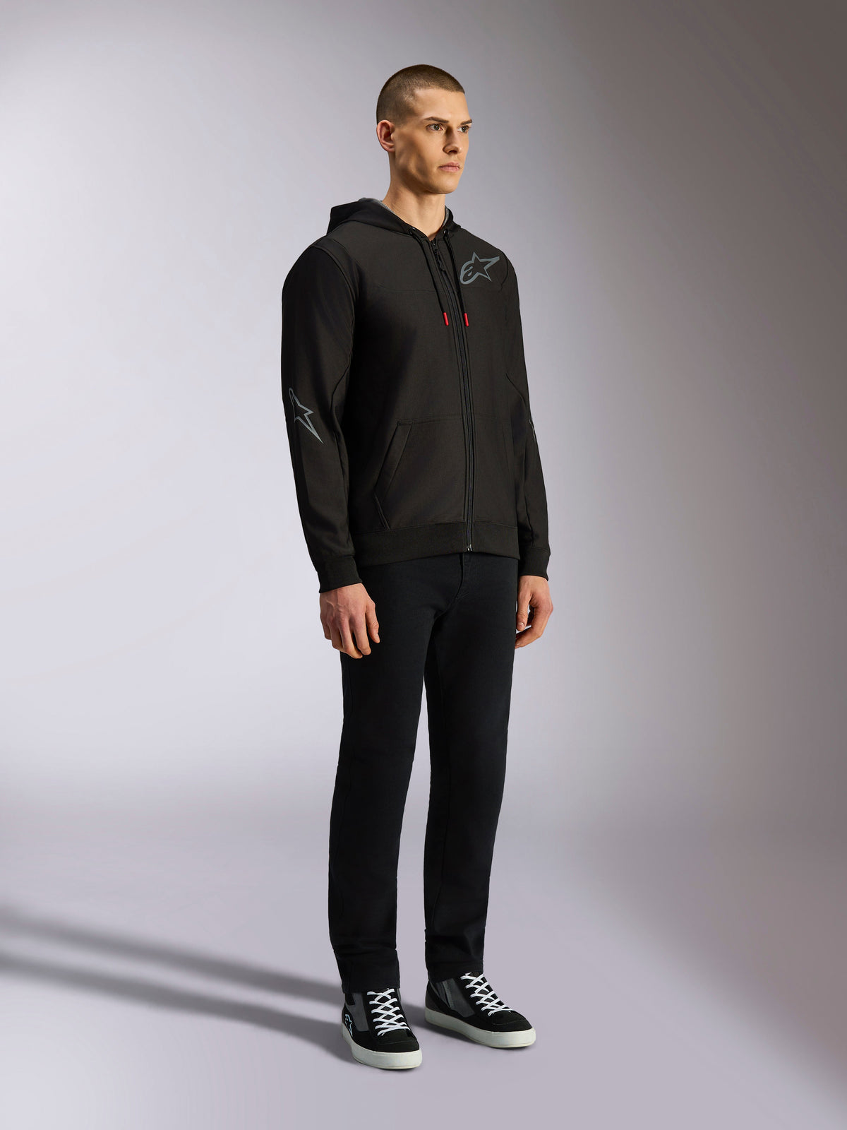 Alpinestars Sessions V3 Hoodie, avslappnad motorsport-streetwear, svart huvtröja med dragkedja, grå stjärnlogotyp på bröstet och ärmen, dragskosnören med röda ändar, ribbade muddar och fåll, bärs av modell med svarta byxor och sneakers.