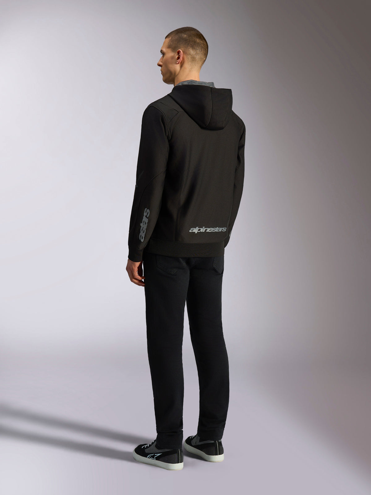 Alpinestars Sessions V3 Huvtröja, vardaglig motorsport-streetwear, svart med grått ordmärke på nedre ryggen och underarmen, polyfleece med dragkedja fram och meshfodrad huva, ryggvy på modell, modern atletisk design